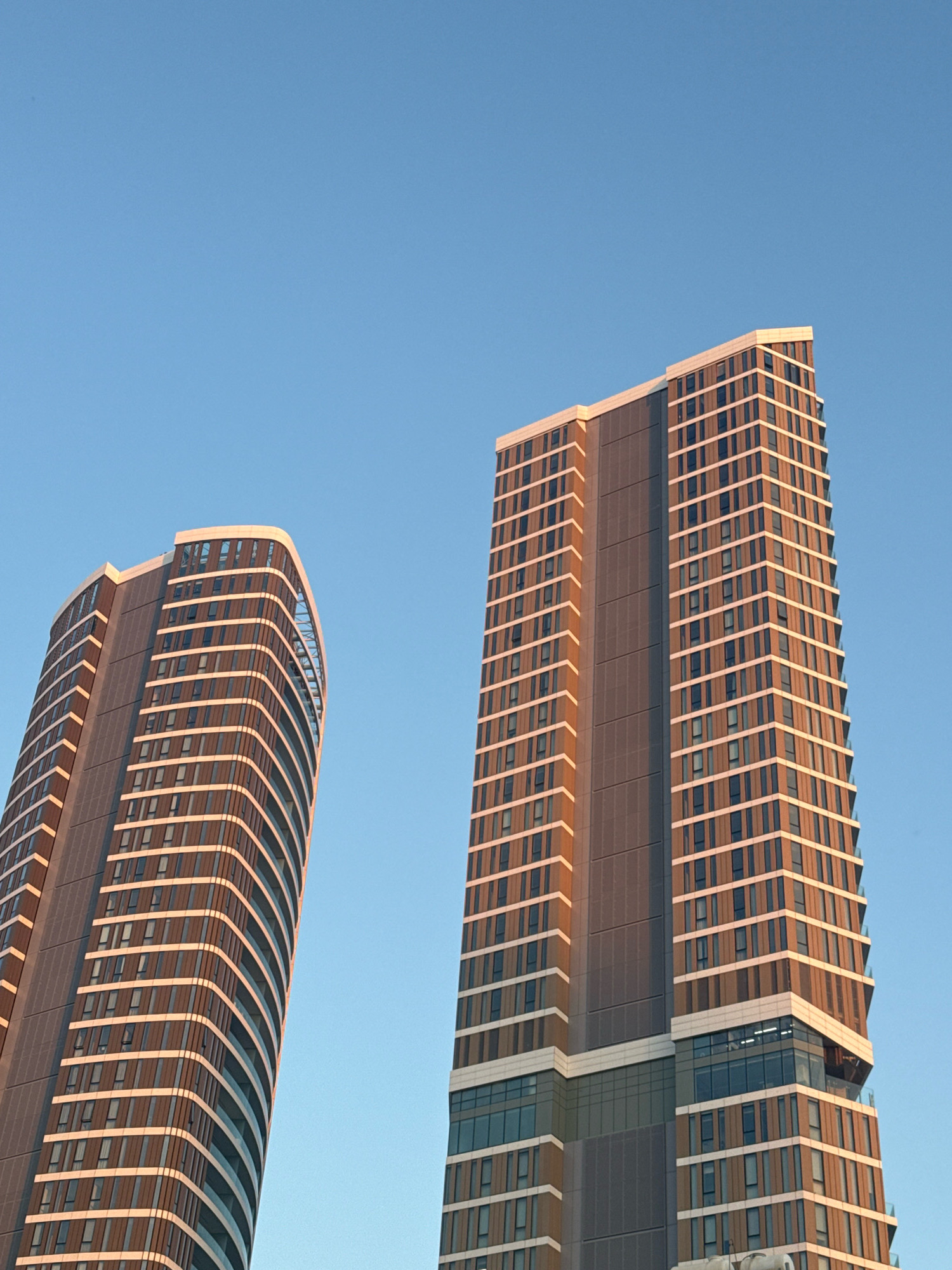 Two modern, high-rise apartment buildings stand against a clear blue sky. The building on the left has a curved facade and is clad in dark gray panels and light-colored horizontal bands, interspersed with numerous windows. The building on the right is taller and more rectangular, with a similar color scheme of dark gray and light bands, also featuring many windows. The late afternoon sun casts a warm glow on the facades of both buildings, highlighting the texture of the materials. The buildings appear to be residential, possibly part of a new development. The style is contemporary urban architecture, typical of rapidly developing city centers. The scene is devoid of people or any specific activity, focusing solely on the architectural structures and their interaction with natural light. The image does not contain any specific landmarks that would definitively place it in Limassol, Cyprus, however, the architectural style is consistent with modern developments in coastal cities.