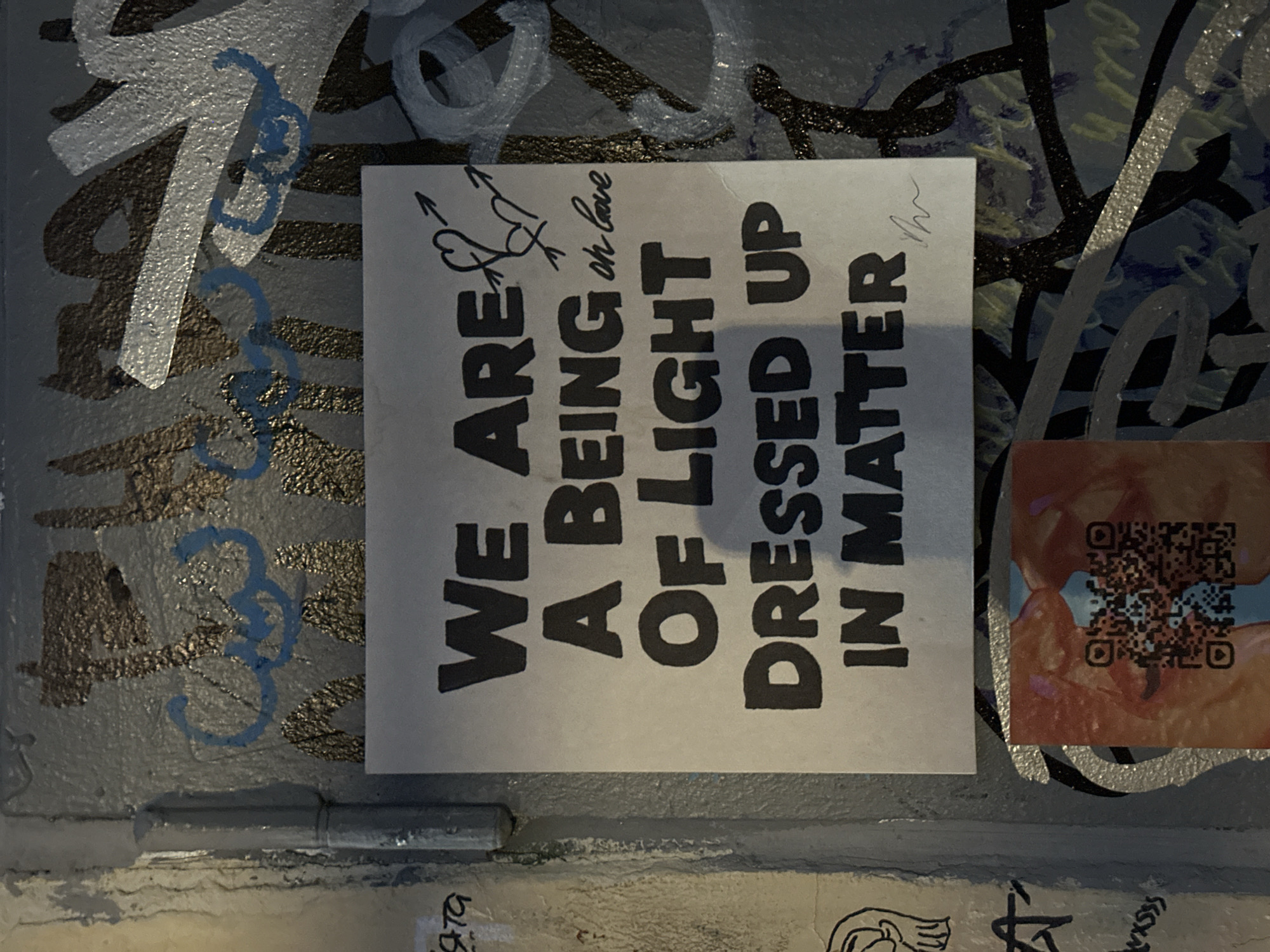 A photograph shows a close-up of a concrete wall covered in graffiti and stickers. Prominently featured is a rectangular sticker with the text "WE ARE A BEING OF LIGHT DRESSED UP IN MATTER." Above this text is a small drawing of two hearts pierced by arrows, along with the words "or love." The sticker is slightly off-white and contrasts with the gray concrete surface. To the right of the sticker is another sticker with an indistinct image and a QR code. The concrete wall surrounding the stickers is adorned with various graffiti markings in silver, blue, black, and other colors. The location is Unknown, Unknown. The scene is unpopulated.