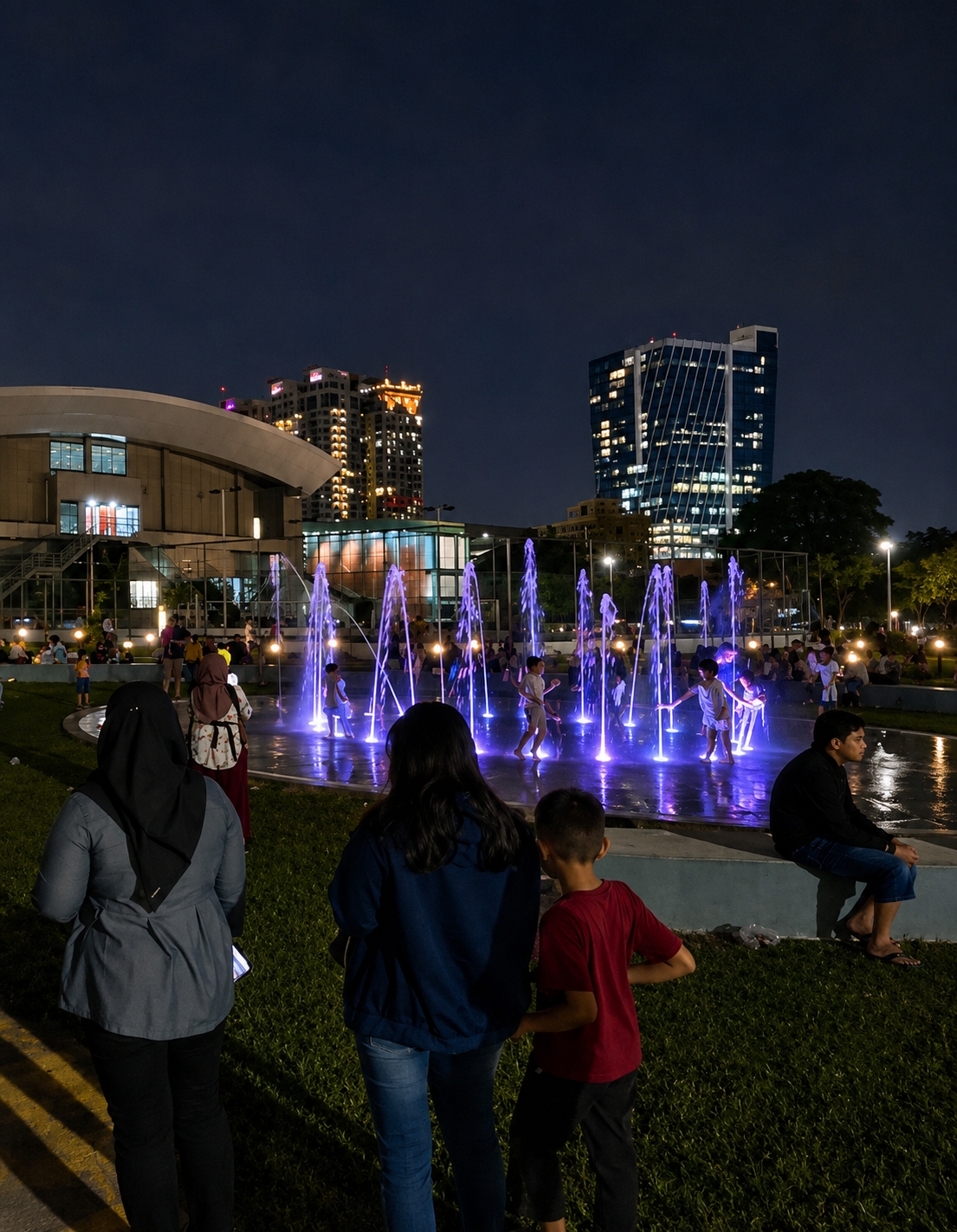 A nighttime view captures a public fountain area with children playing in the illuminated water jets. In the foreground, three individuals are seen from behind: a woman wearing a hijab and a gray jacket, a young woman in a dark blue hoodie, and a young boy in a red t-shirt. Behind them, a man sits on a low wall on the right. The background features modern buildings, some illuminated with numerous windows, hinting at a city environment. The scene is set in the City of Medan, Indonesia, a vibrant urban center known for its blend of modern architecture and public spaces.