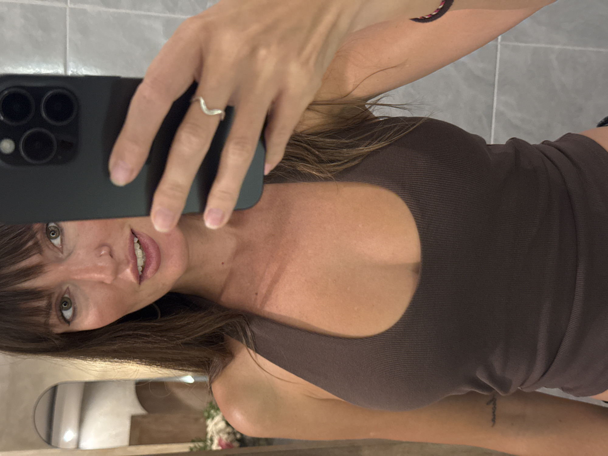 A fair-skinned woman is taking a selfie in a bathroom. She is holding a black smartphone in her left hand, partially obscuring her face. The woman has brown hair with bangs and wears a brown tank top. A silver chevron-shaped ring is visible on her left ring finger. The bathroom features gray tiled walls and a glimpse of a vanity mirror and a floral arrangement is visible in the lower-left corner. A bracelet is on her left wrist and a tattoo is on her right wrist. The location is North Kuta, Indonesia.