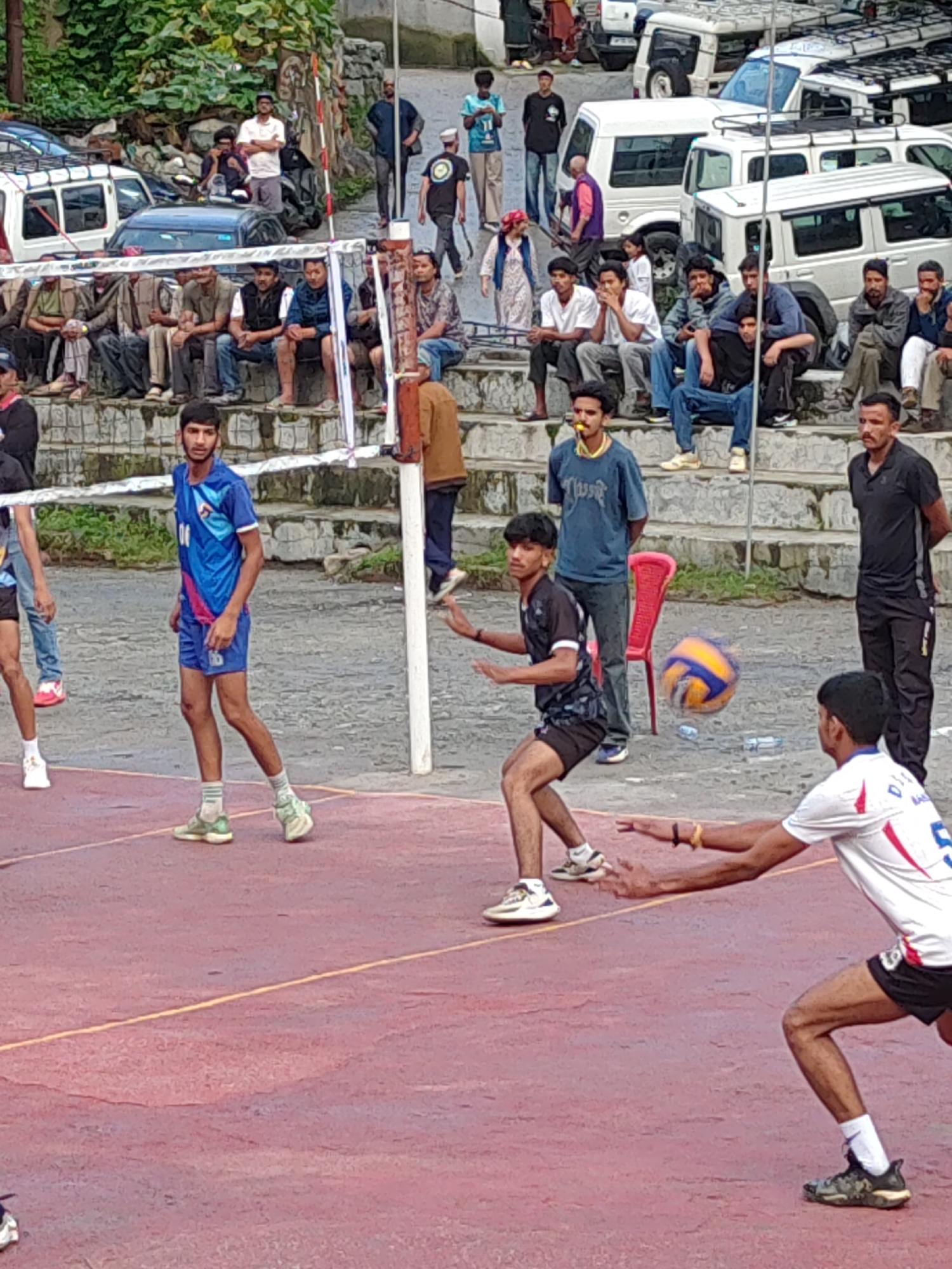 The image captures a volleyball game on a red-colored court, likely in Burwa, India, given the context of previous interactions. Several individuals are actively engaged in the sport, with two men in the foreground positioned to play. One man, wearing a black and white patterned t-shirt and black shorts, is in a defensive stance, his arms outstretched, ready to receive the ball. The volleyball, blurred by motion, is in the air between him and another player. This second player, in a white t-shirt with red and blue accents and black shorts, is also reaching out, appearing to be in the process of hitting or blocking the ball.
Behind these players, a young man in a blue jersey with the number '10' visible, and blue shorts, stands looking towards the game. Further in the background, a group of spectators are seated on tiered stone steps, observing the match. Some are looking at the game, while others are engaged in conversation. A red chair is placed near the spectators.
The environment suggests an outdoor setting with trees and foliage visible in the upper left corner. Several vehicles, including white SUVs and a dark-colored car, are parked in the background, along with what appears to be a building or structures. The overall scene depicts a moment of athletic competition within a community gathering.