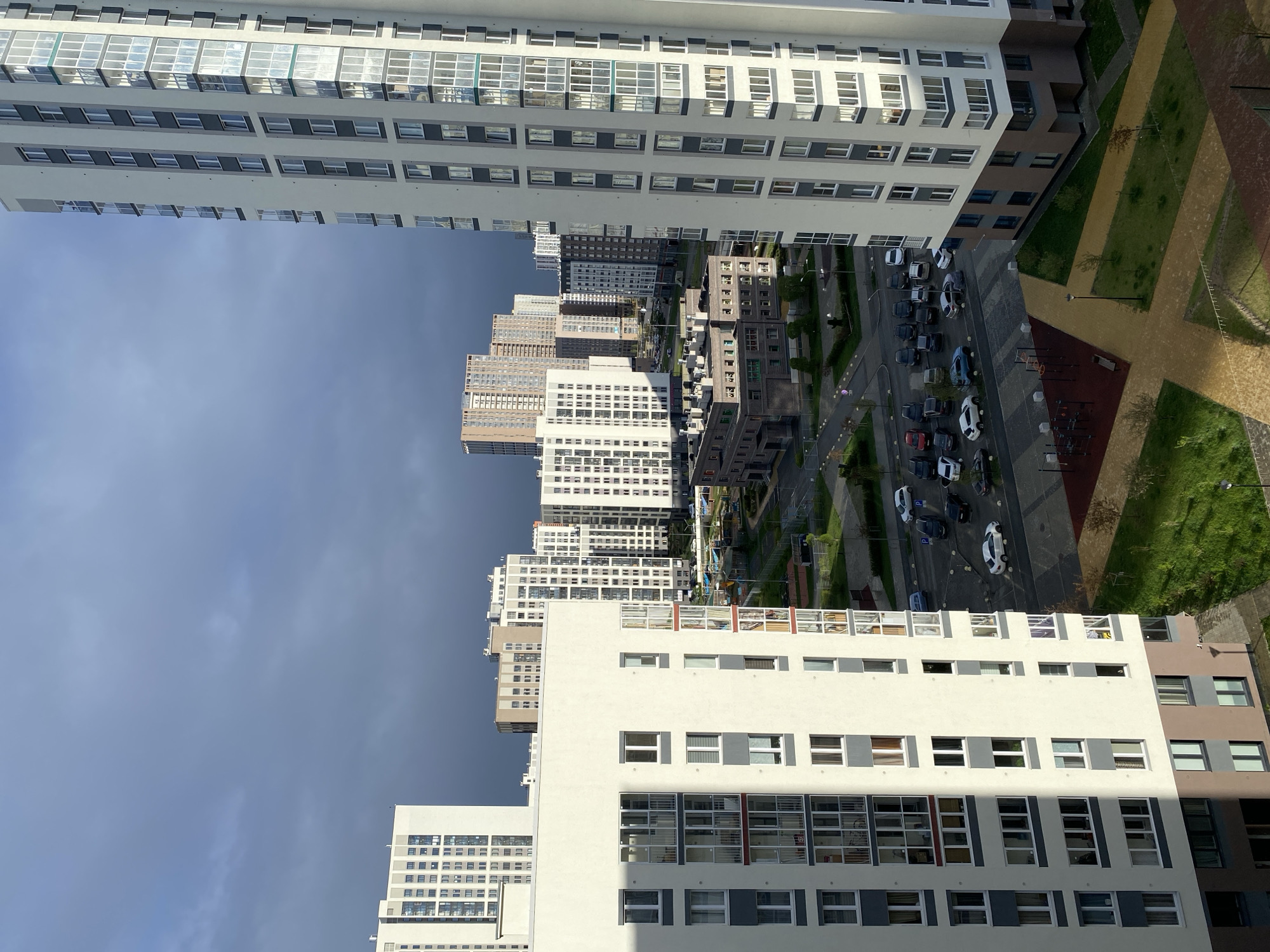 An elevated angle shot shows multiple buildings in what seems to be a dense urban setting, probably a suburb. The sky is visible above the buildings, occupying a significant portion of the image with a dark hue in some areas. The buildings vary in height, color, and architectural design, but most appear to be residential apartments. A road runs between the buildings, and multiple parked cars are visible on the roadsides. An area with trees, grass, and possibly walking paths is seen on the right. The angle and proximity to the buildings give the impression of the shot being taken from another building nearby. The city, Yekaterinburg, Russia, is not discernible from the image alone without additional context or landmarks.