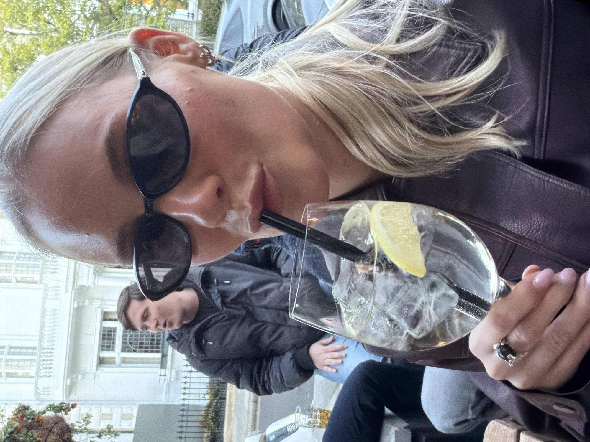 A blonde woman wearing sunglasses and a leather jacket is drinking a beverage with ice and a lemon slice through a straw. A man in a black jacket is partially visible in the background. The location appears to be an outdoor seating area with a view of a white building and a metal fence. The environment suggests an urban setting, possibly in London, UK.