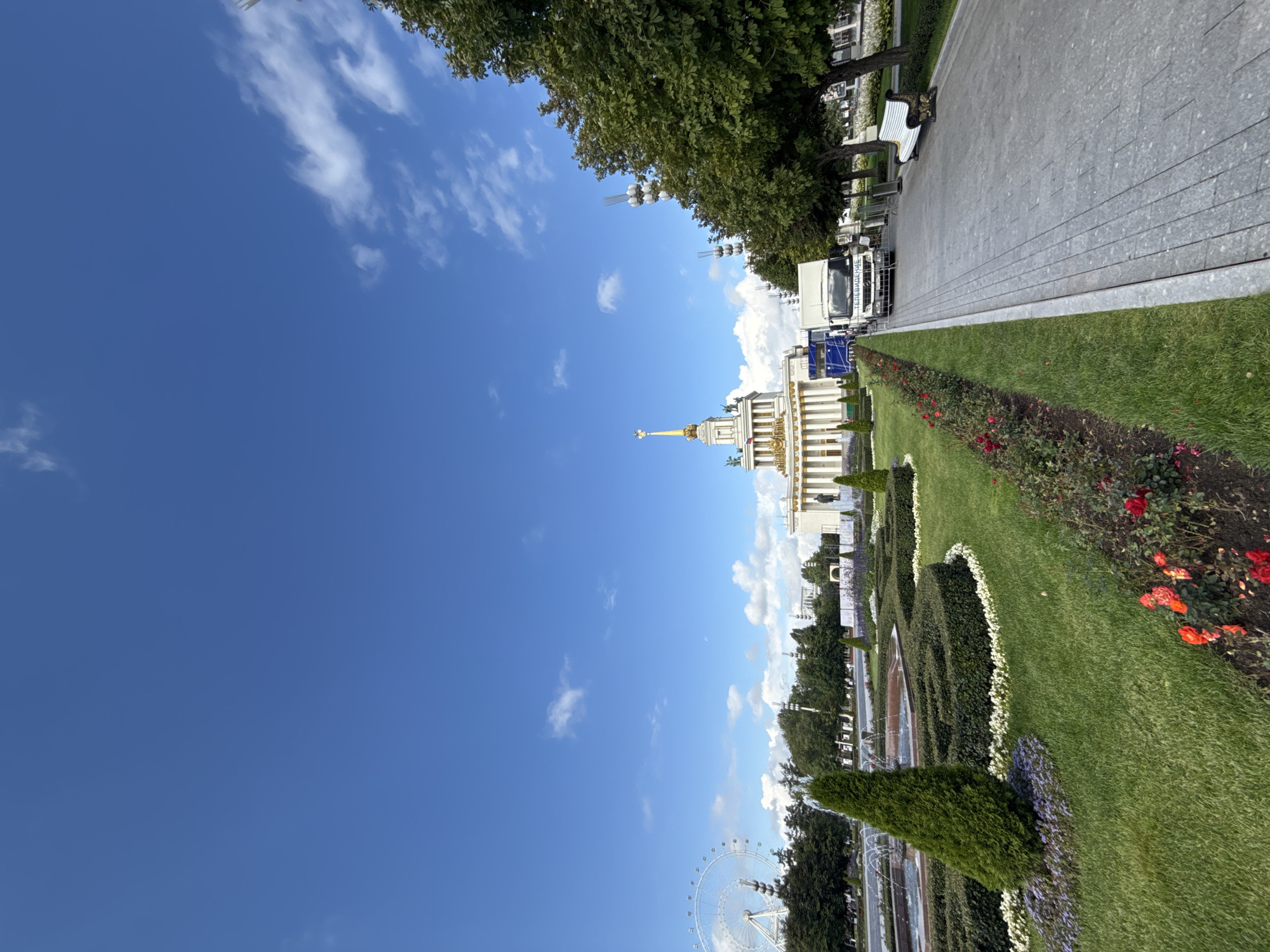 The image depicts a wide view of a park area on a sunny day, with a clear blue sky and scattered white clouds. In the background, a grand building with classical architecture, featuring columns and a golden spire, is visible. To the left of this building, a large Ferris wheel can be seen. The foreground of the image showcases a well-maintained park with lush green lawns, manicured hedges forming geometric patterns, flower beds with red roses and purple flowers, and a fountain. A paved walkway runs along the right side of the park, featuring a white bench with ornate detailing. A white truck with a blue sign is parked near the building. Several trees line the walkway and the park area. The scene appears to be VDNKh (Exhibition of Achievements of National Economy) in Moscow, Russia, a popular park and exhibition center.