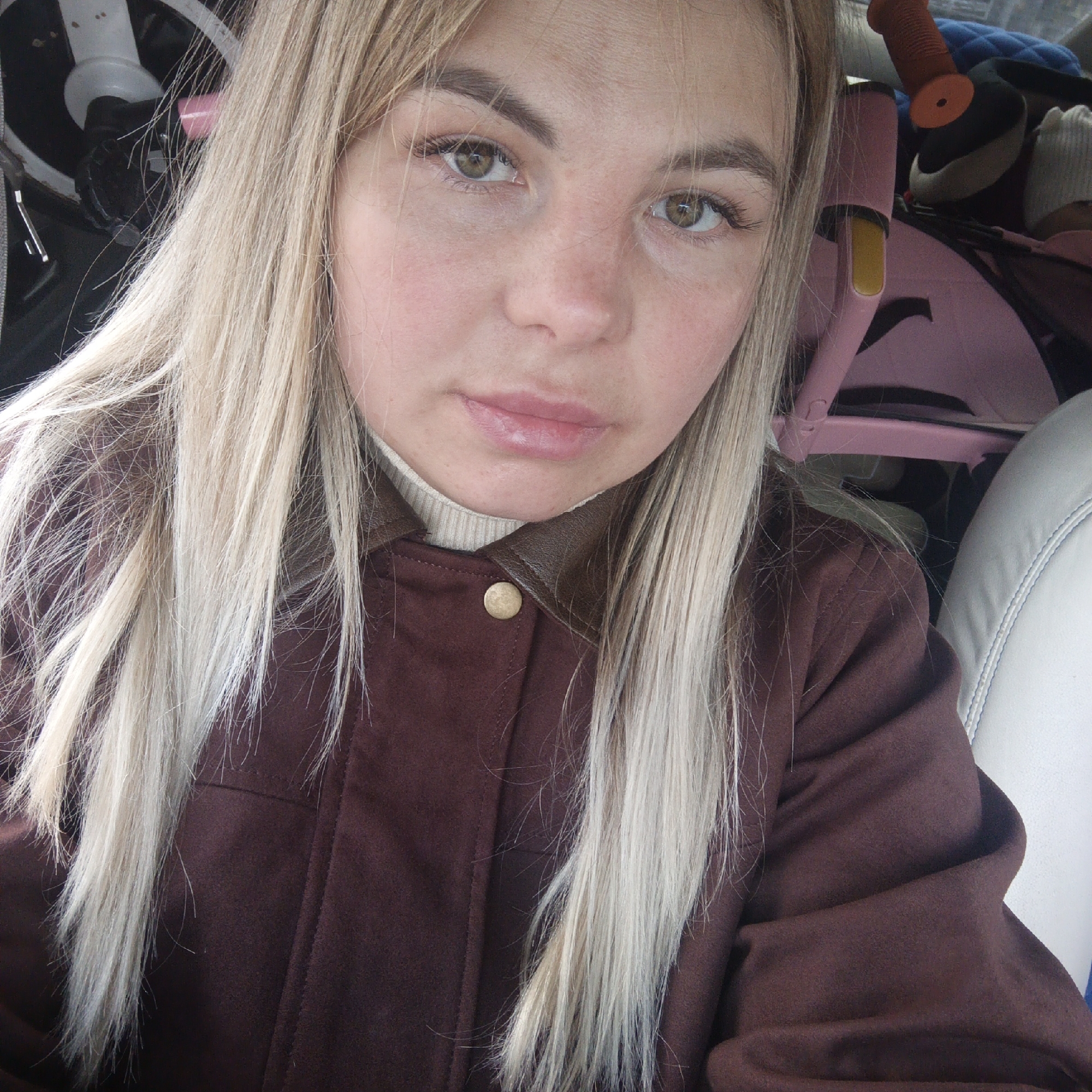 A woman with long blonde hair and a brown jacket is sitting in a vehicle. Her hair is parted in the middle and falls over her shoulders. She is wearing a brown jacket with a leather collar and a button at the neck. Behind her, there is a white seat, a pink child's seat, and other items that are out of focus. The setting is a vehicle interior in the city of Unknown, Unknown.