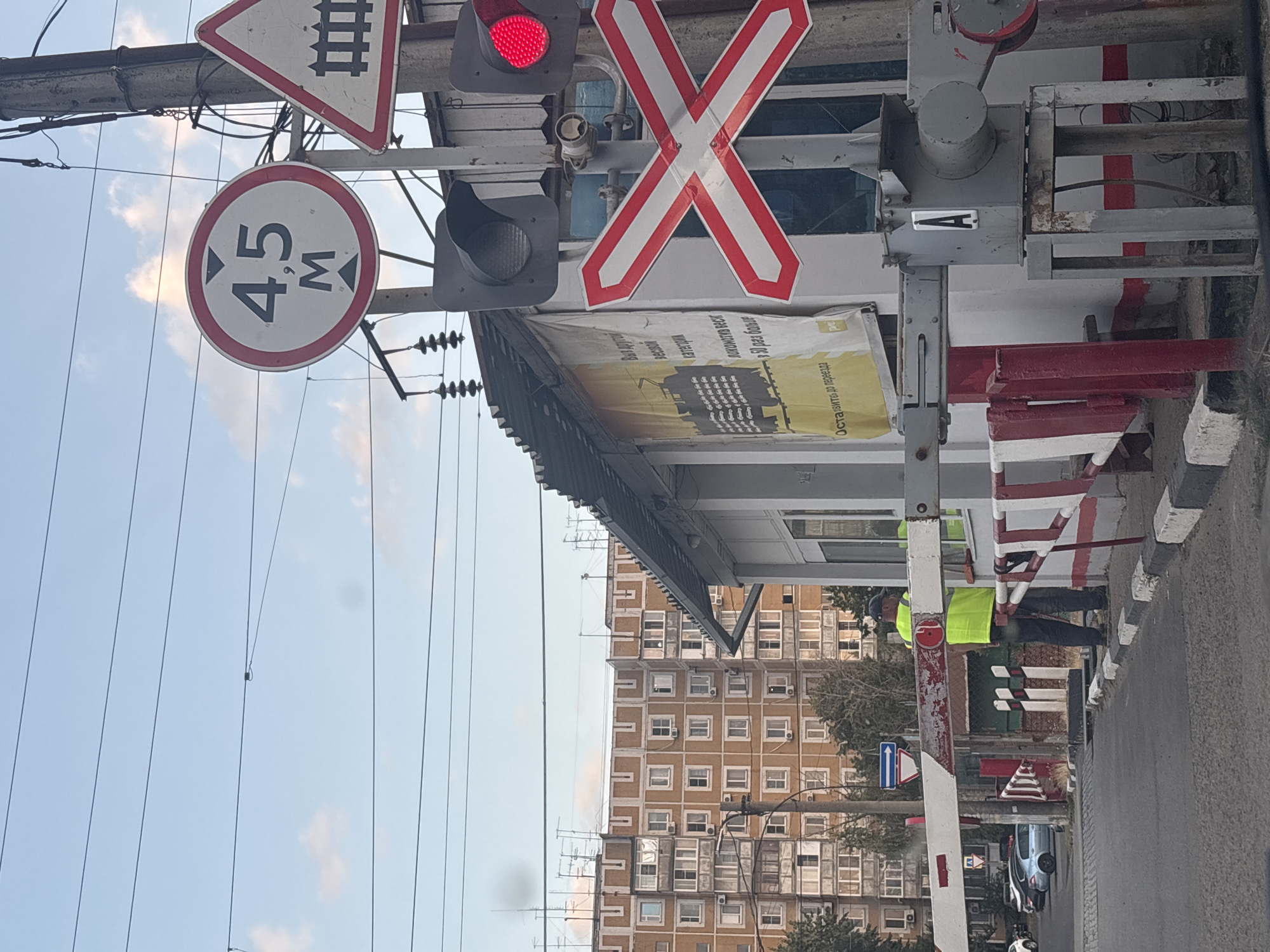 The image depicts a railway crossing in Krasnodar, Russia. A railroad crossing sign, a red traffic light, and a barrier arm are visible. A person wearing a yellow vest stands near the barrier, seemingly attending to the crossing. In the background, a multi-story apartment building and a portion of a street with parked cars are visible, indicating an urban setting. The sky is partly cloudy, suggesting daytime. The overall scene suggests a functional railway crossing within a residential area of the city.