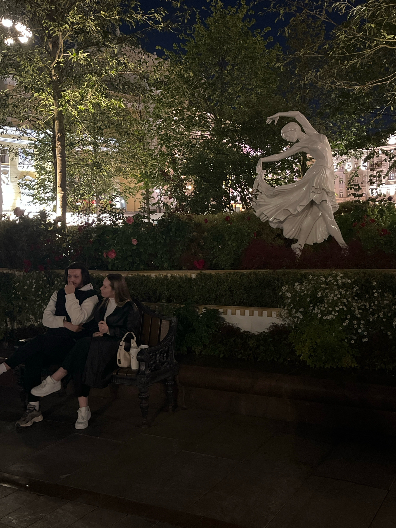 The image depicts a night scene in a park-like setting, possibly in Moscow, Russia, featuring a white statue of a dancer in mid-pose. In the foreground, a young couple is seated on a dark-colored bench. The man, on the left, wears a black vest over a white top and dark pants, with one leg crossed over the other. The woman, on the right, is dressed in a black jacket and dark pants, with white sneakers. A beige handbag rests on the bench beside her. The background is filled with lush greenery, including trees with illuminated foliage and bushes with red roses and white flowers. The lighting suggests the presence of street lamps or architectural lighting, casting a warm glow on the scene. In the distance, blurred buildings with illuminated windows are visible.