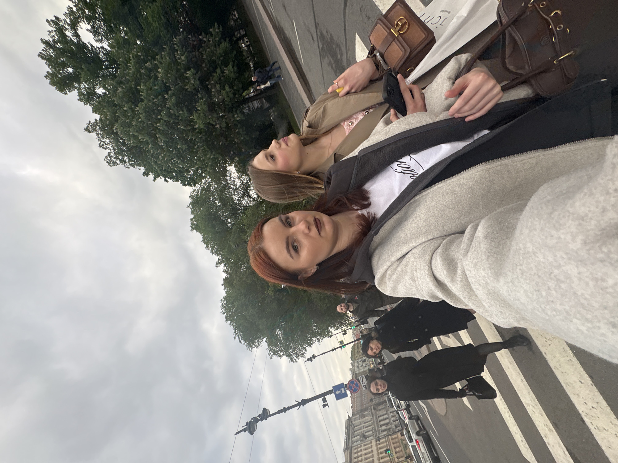 Two women stand closely together taking a selfie. The woman in the foreground has red hair and is wearing a gray hoodie. The woman in the background has brown hair and is wearing a tan jacket. Both are holding brown purses. Behind them, three people dressed in black are crossing a zebra crossing. In the background are buildings, trees, and a cloudy sky. The location is Saint Petersburg, Russia.