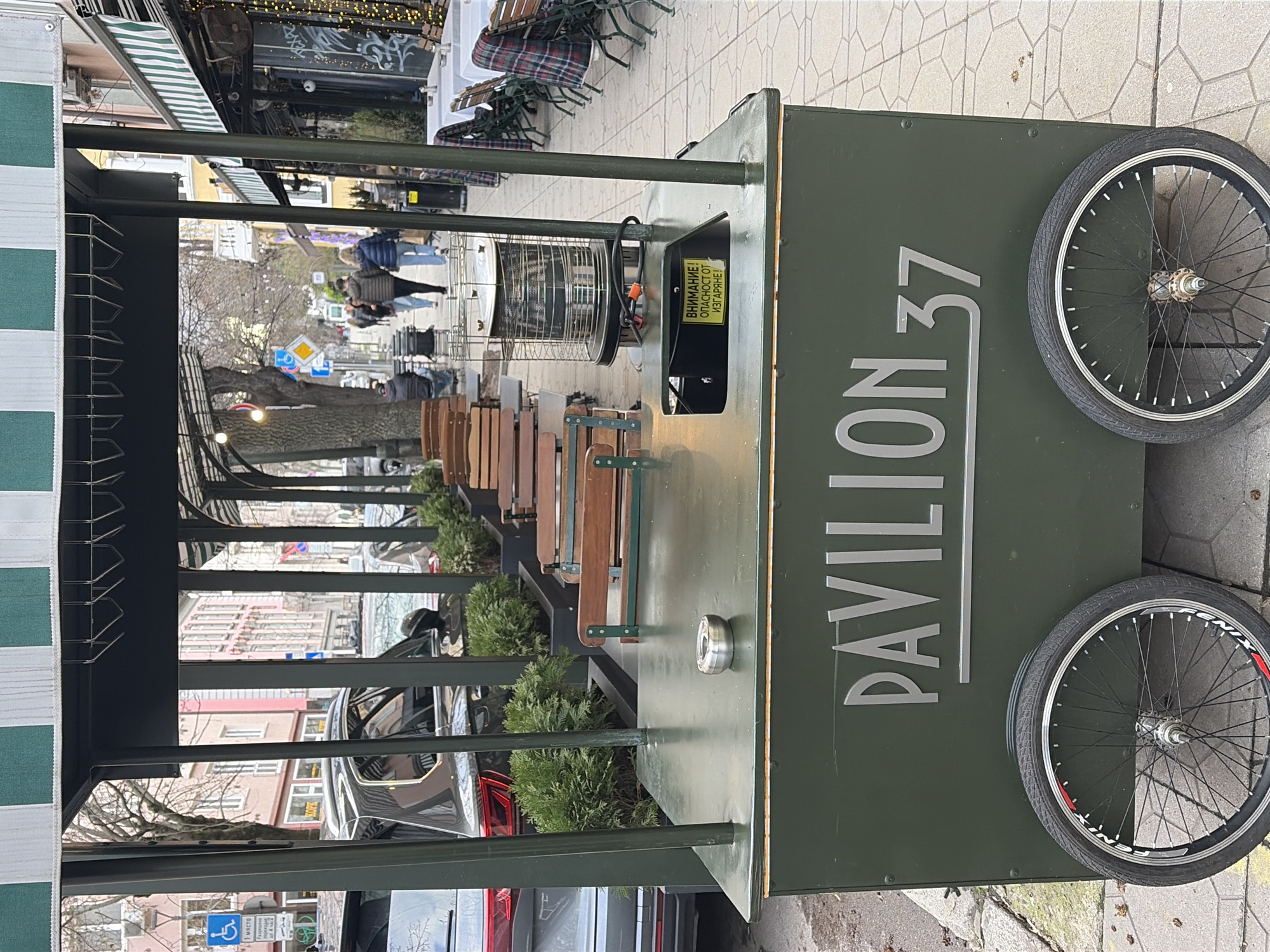 The image shows a street scene in Sofia, Bulgaria, featuring a mobile kiosk or cart labeled "Pavilion 37." The kiosk is painted green and has two bicycle wheels attached for mobility. It is positioned along a sidewalk with outdoor seating for a cafe or restaurant. People are walking in the background, and several cars are parked nearby. The weather appears to be overcast.