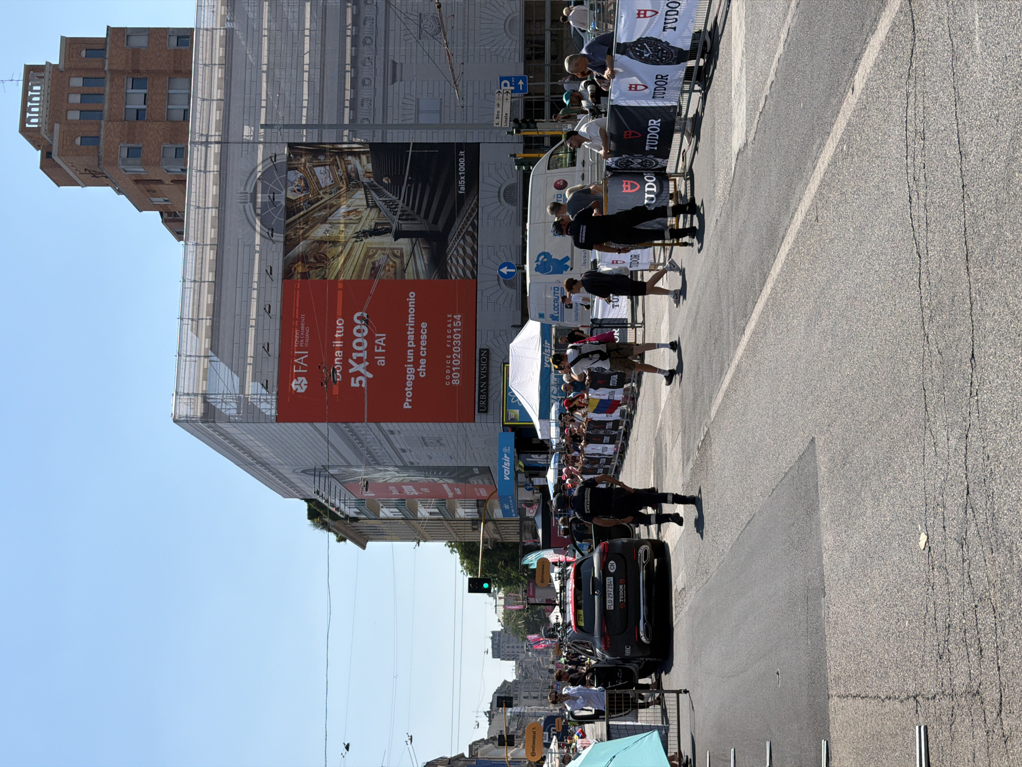 Here is a description of the image:
The image shows a street scene in Milan, Italy, with a cycling event taking place. The event is likely the Giro d'Italia, indicated by the presence of Tudor banners, race cars, and officials lining the route. There's a black car with the license plate "LU-297284 CH" marked with "TUDOR" and "CARABINIERI". A building undergoing renovation is behind the race. It is covered in scaffolding and has a large banner ad for "FAI", an Italian environmental fund, promoting their "5X1000" donation program. The intersection shows a green traffic light. People are standing along the route to watch the race. In the background, the city skyline is visible under a clear, blue sky.