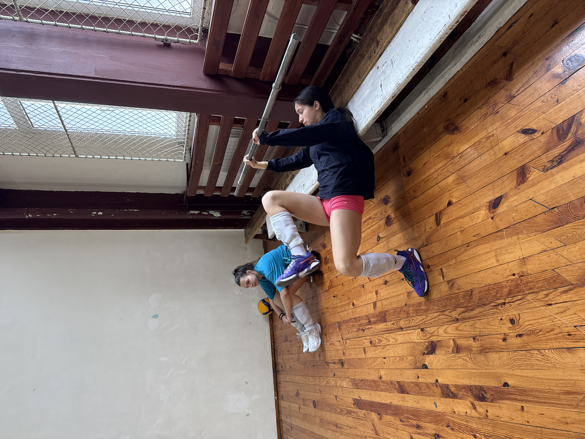 Two women are in a large room with a wooden floor. The room is indoors and has light-colored walls. The woman in the foreground is wearing a black long-sleeved shirt and pink shorts, and she has one leg lifted. She is holding a gray pole above her head. The woman in the background is wearing a blue shirt and has a volleyball. She is bending over. The room has beams and pipes visible in the ceiling, and there is a window covered with netting. The location is likely an indoor sports facility. The city of Sofia, Bulgaria is not discernible from the visual data.