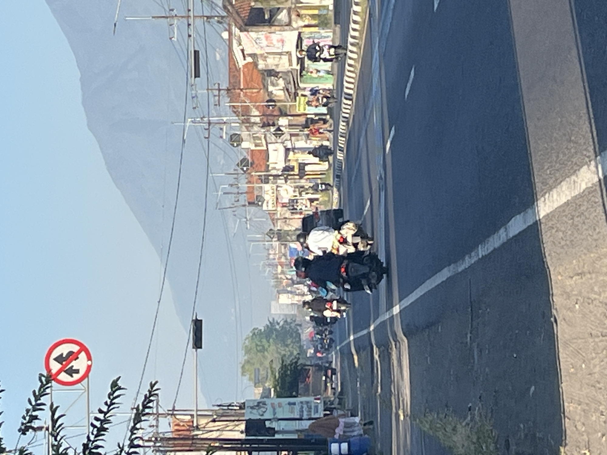 A street scene in Siring, Indonesia, is captured under a clear sky. A mountain range is visible in the background. A traffic sign prohibiting the overtaking of motorcycles is posted on the left side of the image. The road is busy with traffic, primarily motorcycles, with a few cars present. Buildings line the right side of the road, some of which appear to be shops or homes. The road surface shows signs of wear and tear, with some unevenness and cracks. The scene depicts a typical day on a busy Indonesian street.