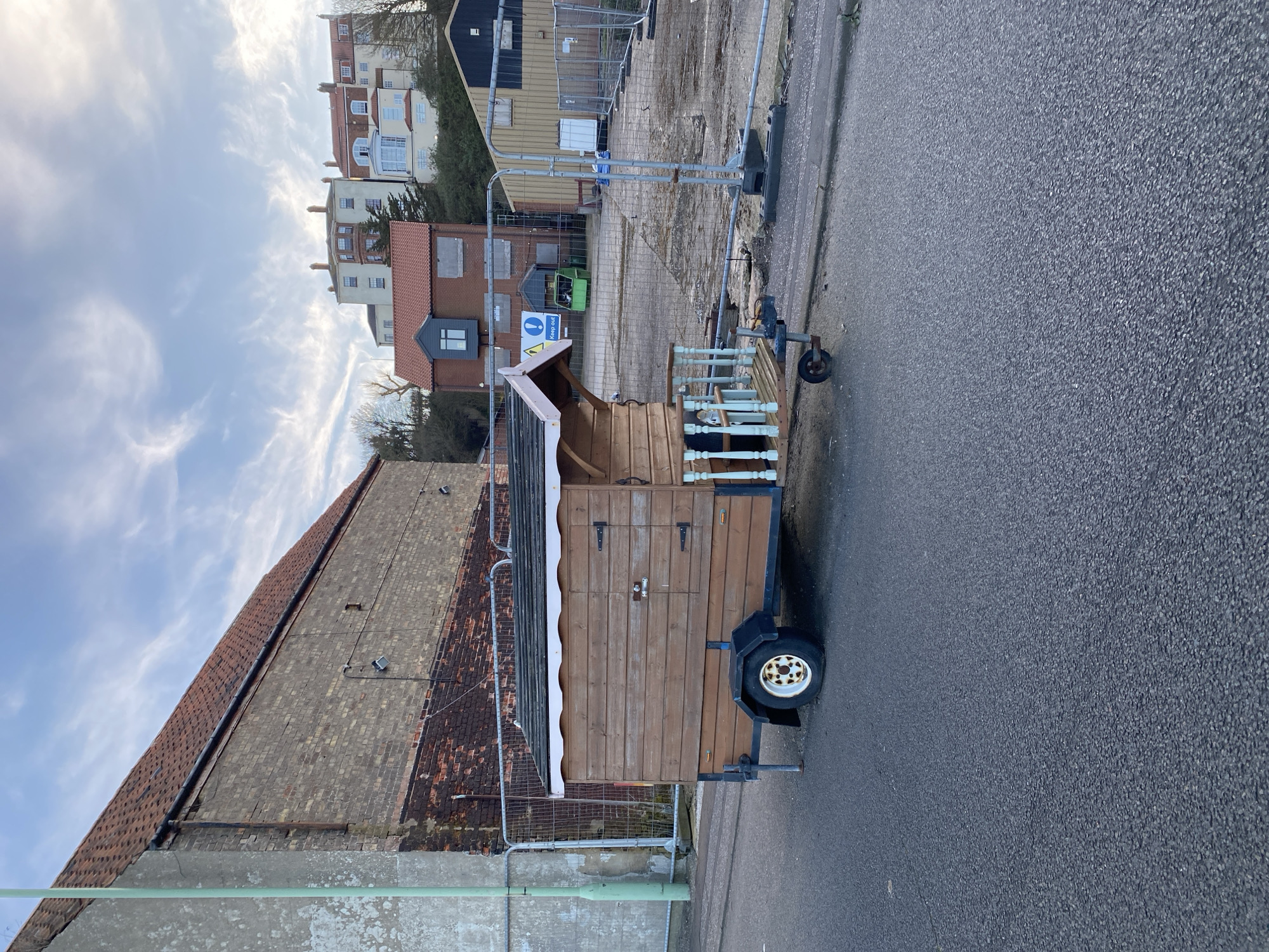 A wooden shed-like structure on a wheeled trailer sits on an asphalt road adjacent to a weathered brick building with a pitched roof. A temporary metal barrier separates the structure from the building. In the background, a construction site enclosed by chain-link fencing is visible, with a multi-story building further back. The sky is partly cloudy with a blue hue. The location is in East Suffolk, United Kingdom.