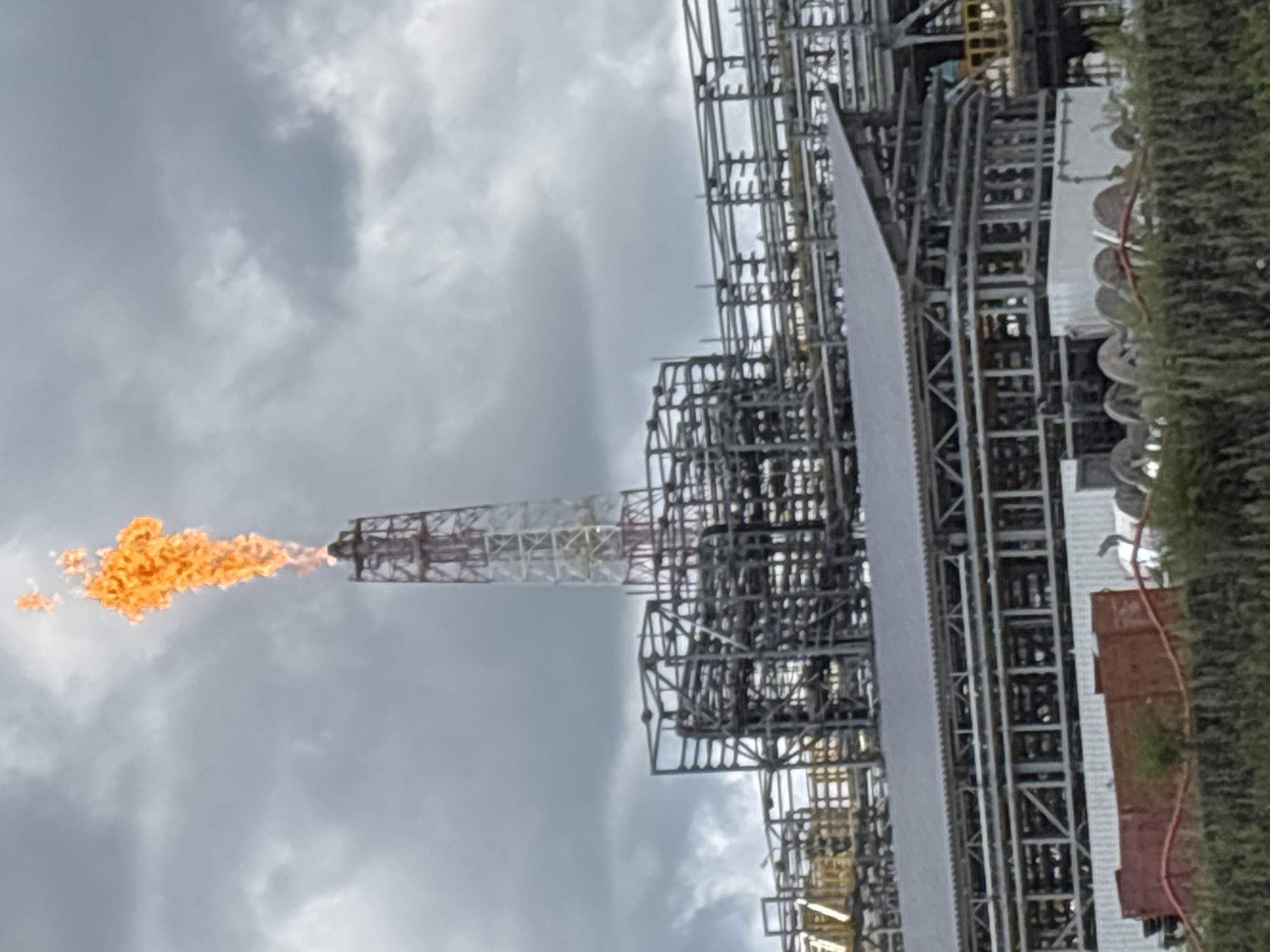 An industrial flare stack is visible in Chernigovka, Russia, spewing bright orange flames high into a cloudy sky. The flare stack, a tall, lattice-like structure with red and white markings, is part of a larger industrial complex comprised of extensive pipework and metal structures. Below the flare stack and complex, a red industrial bin and green foliage are visible. The overall scene suggests an active industrial facility.
