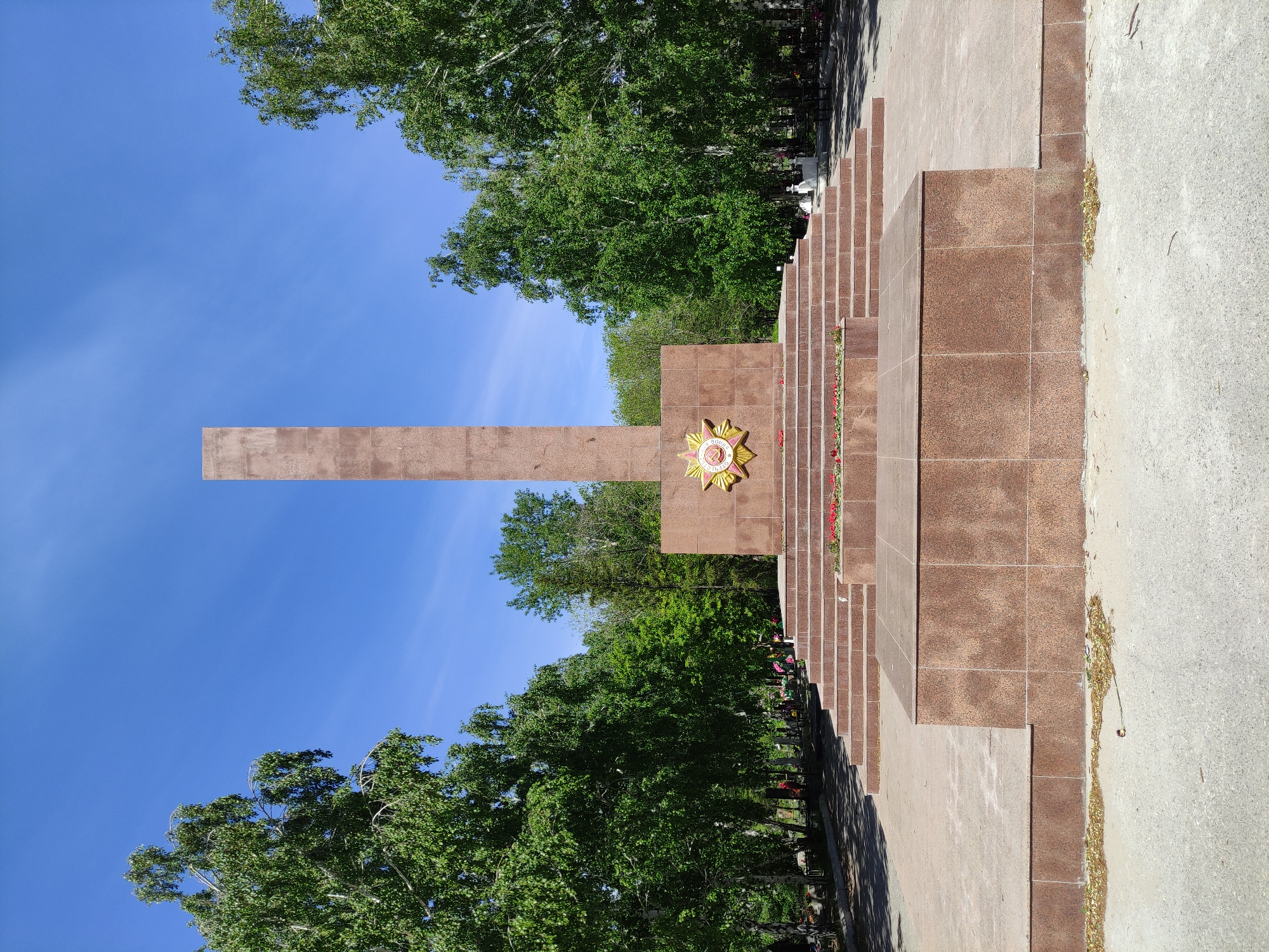 The image shows a monument in Pavlodar, Kazakhstan, dedicated to the veterans of the Great Patriotic War. The monument is made of brown stone. It consists of a vertical slab intersecting a horizontal rectangular base. A Soviet-era star emblem adorns the face of the monument, and red flowers are laid at its base. A series of steps lead up to the monument. The monument is located near trees and a cemetery. The sky is visible in the background.