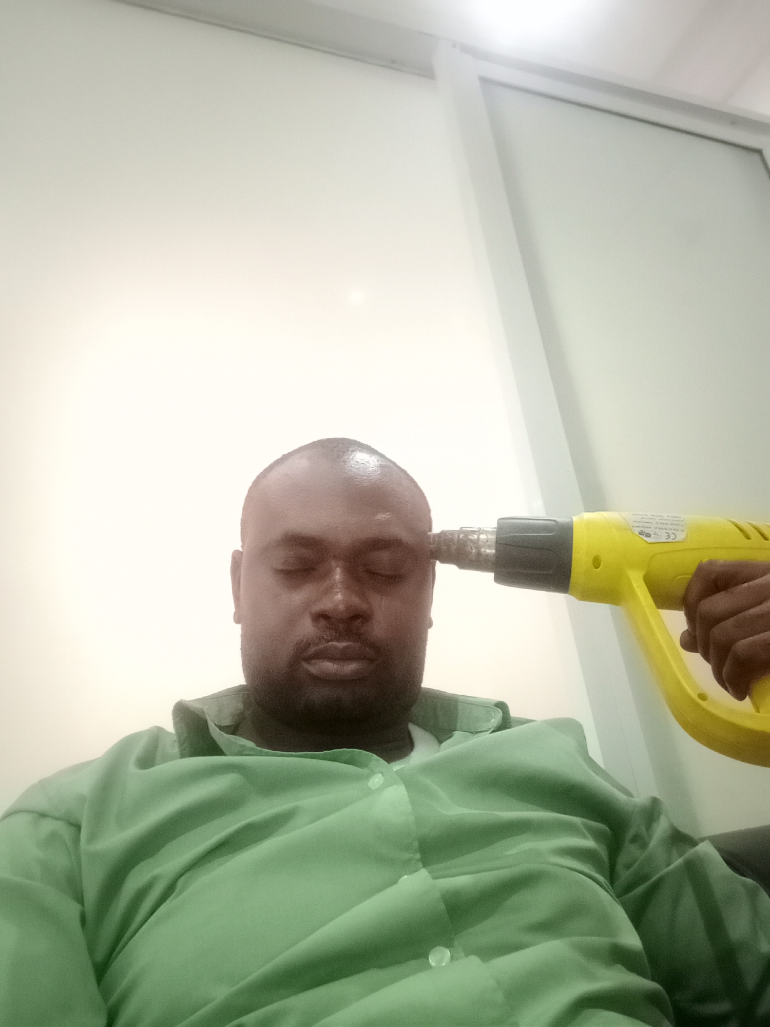 An indoor, close-up shot features a dark-skinned man with a bald head and a short beard, wearing a light green collared shirt. His eyes are closed, and he maintains a neutral expression, possibly indicating relaxation or a dramatic pose. A bright yellow industrial heat gun is positioned with its nozzle against his right temple, held by a dark-skinned hand.
The setting appears to be a brightly lit room with plain white walls. A white door frame or partition is visible in the background on the right side of the image. The lighting is bright, with a soft glow in the upper left corner. On the side of the yellow heat gun, small labels and text are visible, including a clear "CE" mark.
The overall scene suggests a humorous or staged moment, perhaps playfully depicting stress or "blowing one's mind" using an unexpected object like a heat gun instead of a firearm. The image provides no specific time of day but appears to be well-lit indoors. This photo was taken in Gwarinpa, Nigeria.