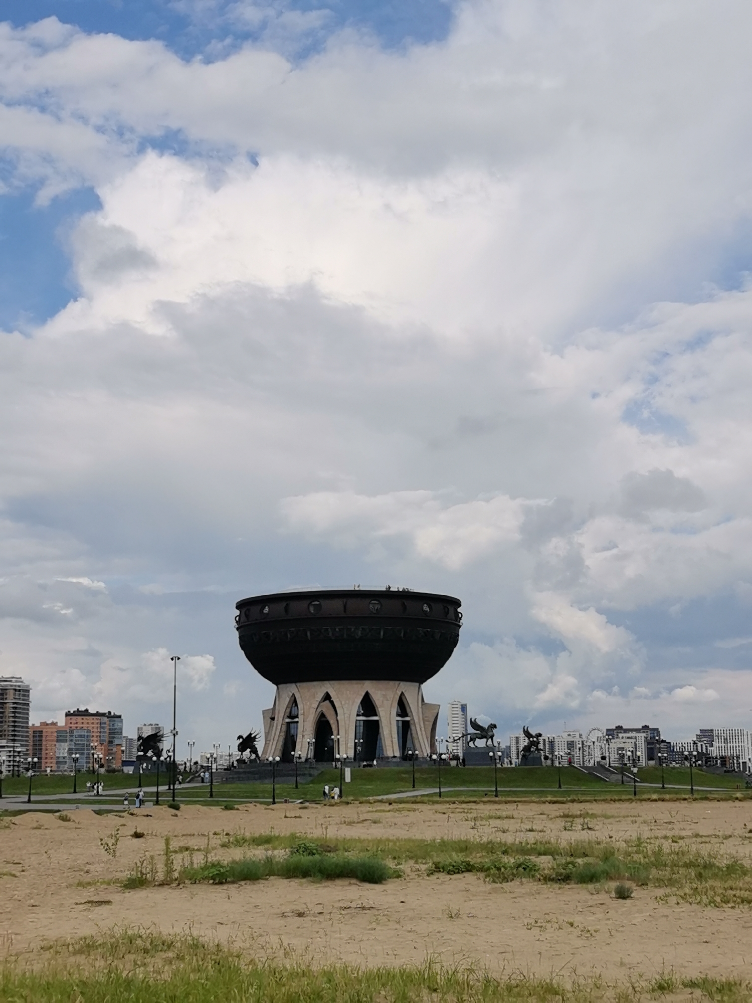 The image depicts the Family Center "Kazan" in Kazan, Russia, also known as the "Chalice" due to its distinctive cauldron-like shape. The structure, a prominent landmark, stands against a cloudy blue sky. The building features a large, dark, rounded upper section supported by several arching structures. In front of the building, a grassy area is visible, with paths and lampposts. Two winged mythical creatures, possibly griffins or sphinxes, are mounted on pedestals on either side of the entrance. In the background, modern high-rise buildings are visible, forming the city skyline. The foreground is a sandy area with sparse green grass, suggesting a park or undeveloped land. Several people are scattered throughout the scene, walking and observing the landmark. The overall impression is a panoramic view of an architectural monument and its surrounding urban landscape.