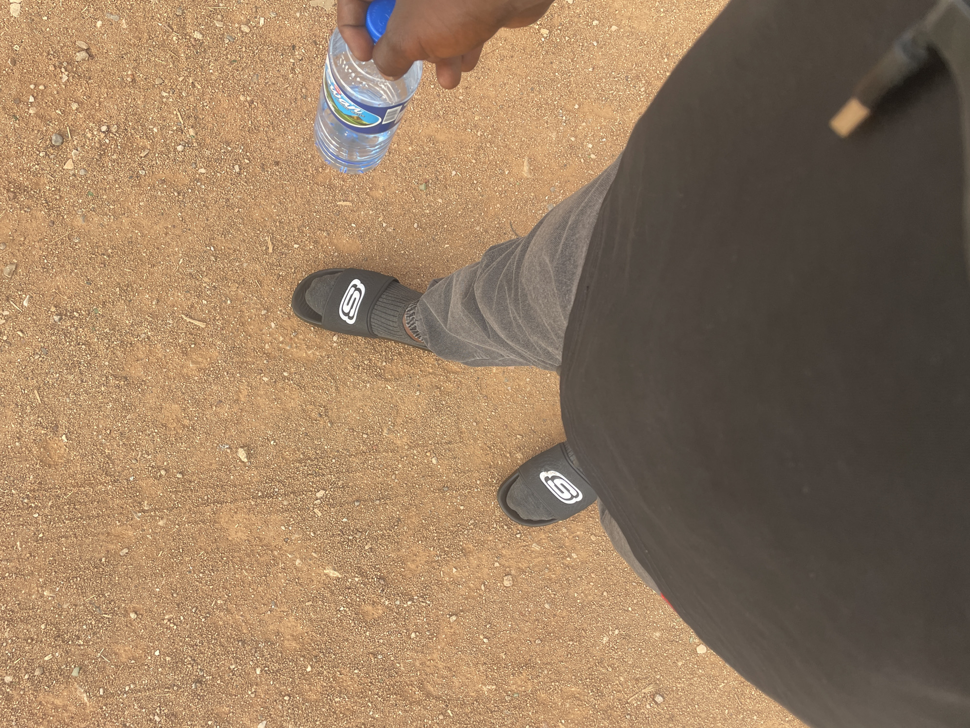 The image is a top-down view showing a person standing on a dirt or gravel surface in Jos, Nigeria. The individual is holding a plastic bottle of water. They are wearing grey pants and black socks and Skechers slides. A black bag or backpack is also visible. The ground is composed of small, brown particles.