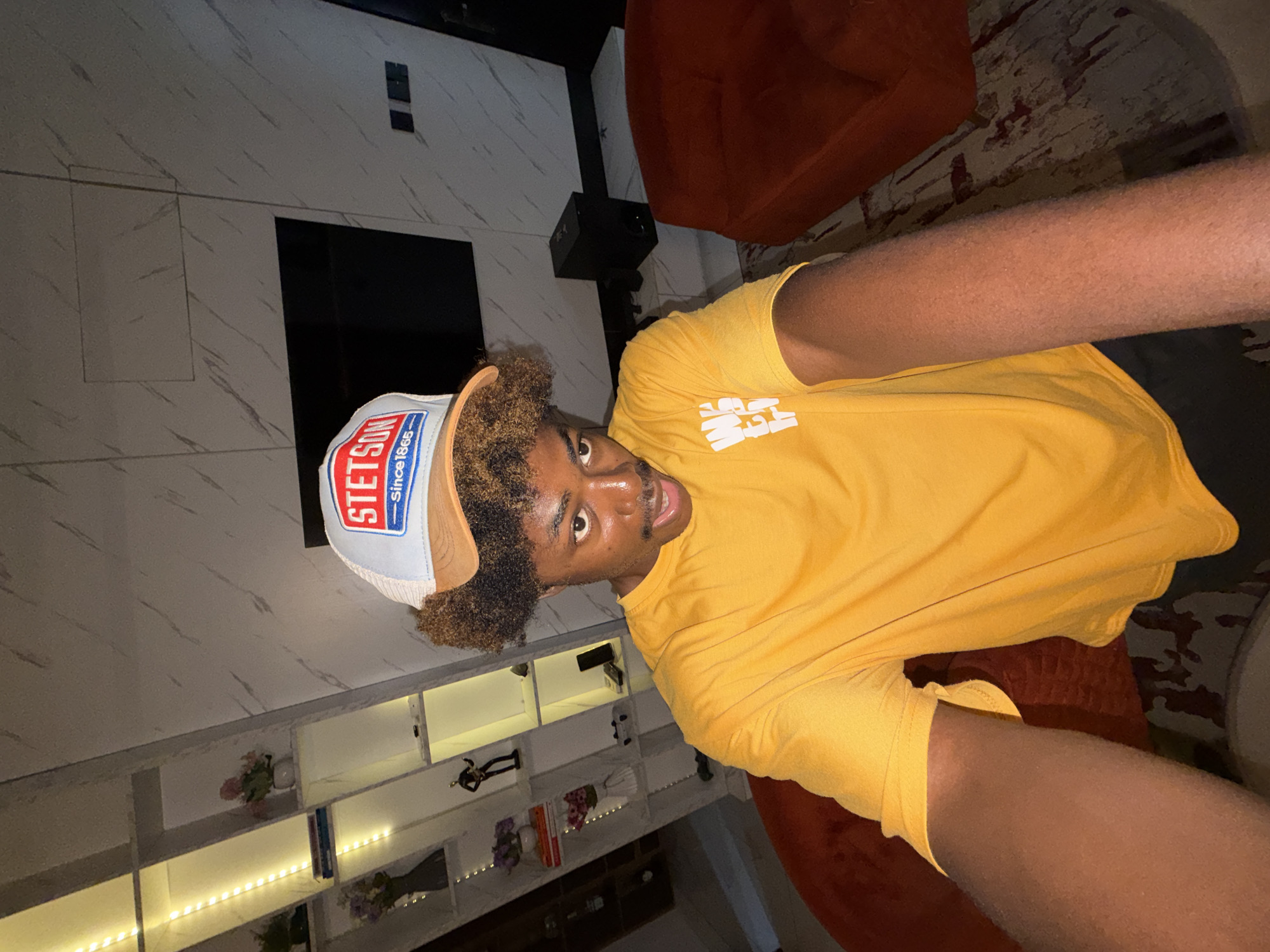 A man is lying on a patterned carpet in a room that appears to be in a modern home. He is wearing a yellow t-shirt and a Stetson-branded baseball cap. The background includes a light grey wall with marble-like patterns and a built-in entertainment center with shelves and decorative items. There is a dark TV screen on the wall. There's also what looks like a speaker mounted near the top of the wall. The room includes modern furniture such as a red beanbag. Based on the location provided, the scene is presumably in Igboefon, Nigeria.