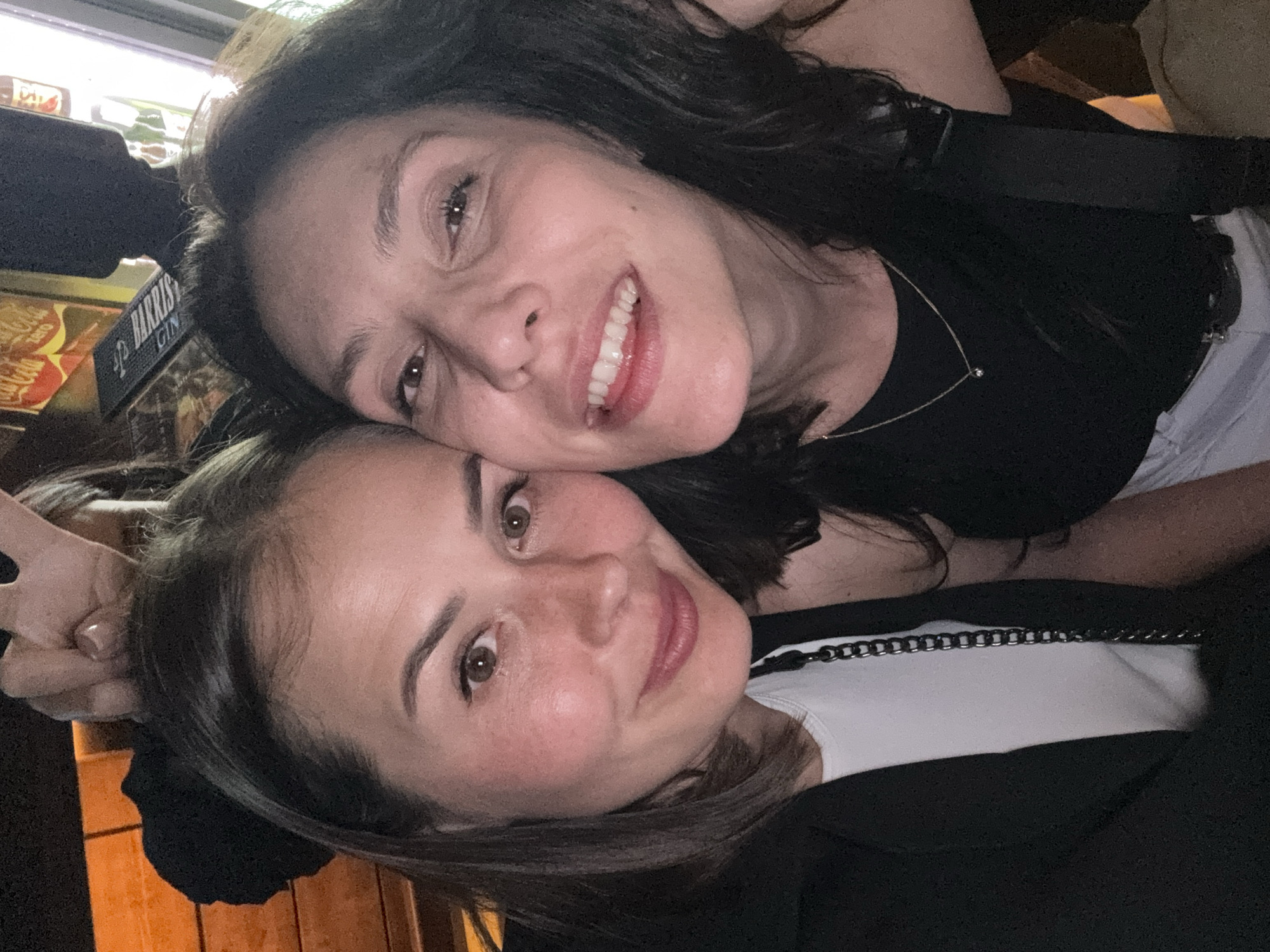 Two women are posing closely together for a selfie-style photograph. The woman on the top has dark, wavy hair, fair skin, and is wearing a black top with a delicate necklace. Her expression is a bright smile with her teeth visible. The woman on the bottom has dark hair, fair skin, and is wearing a white top under a dark blazer. She is making a peace sign with her hand, and smiling at the camera. In the background, there's a partially visible Coca-Cola sign, and what appears to be a HARRIS Gin bar sign. Given the presence of the signs, the scene appears to be inside a bar or restaurant. It is impossible to confirm the location in the city of Chaikovsky, Russia from the provided image and information.