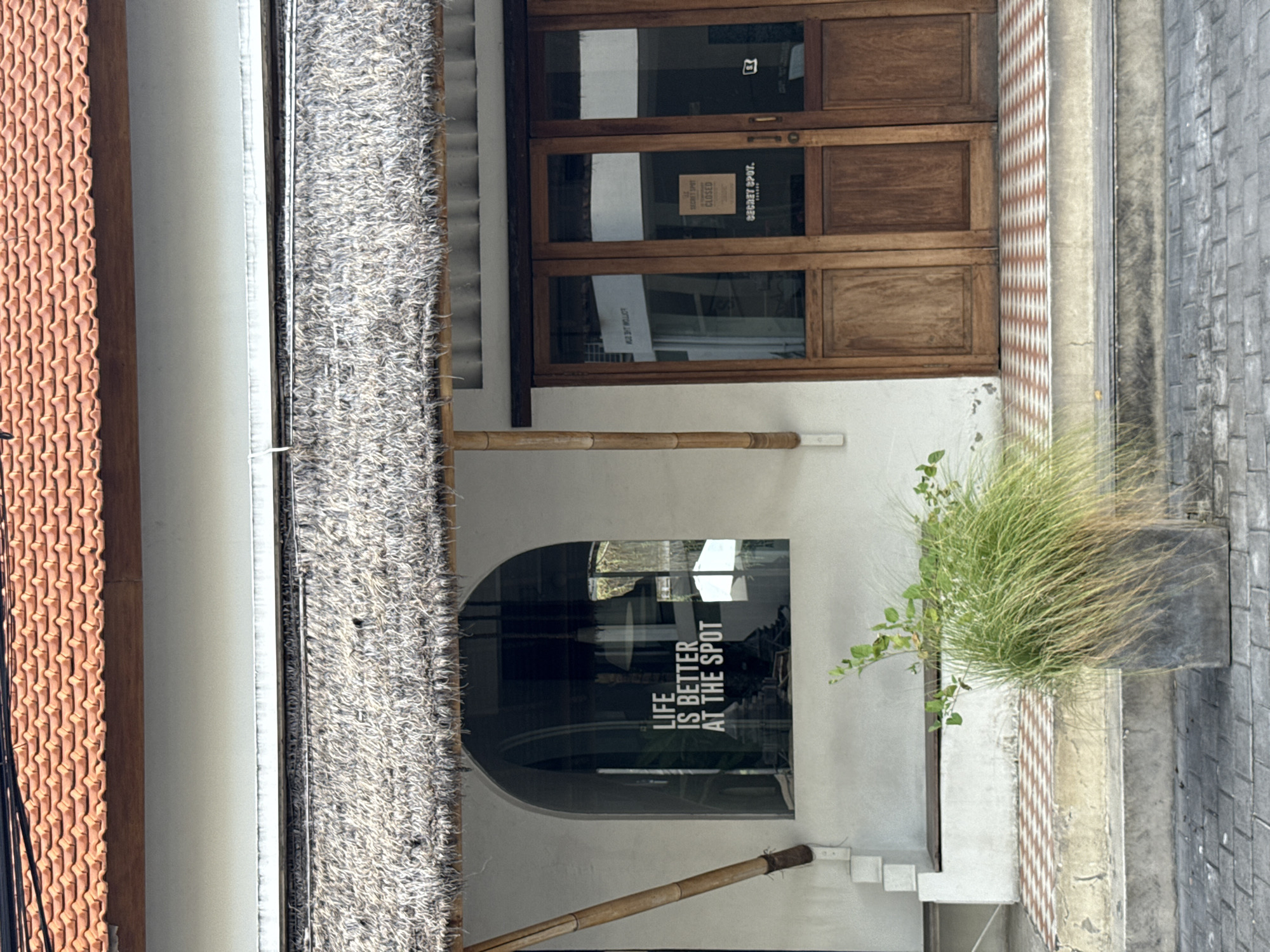 The image depicts the exterior of "Secret Spot" in North Kuta, Indonesia. The structure has a red tile roof on the left and a thatched awning extending over the façade. The entrance features a set of wooden doors with a "CLOSED" sign visible. Below the doors, there is a white wall with a rounded window. Inside the window, text reads "LIFE IS BETTER AT THE SPOT". A bamboo support structure runs horizontally along the wall. To the right of the window, a potted plant sits near a tiled sidewalk.
