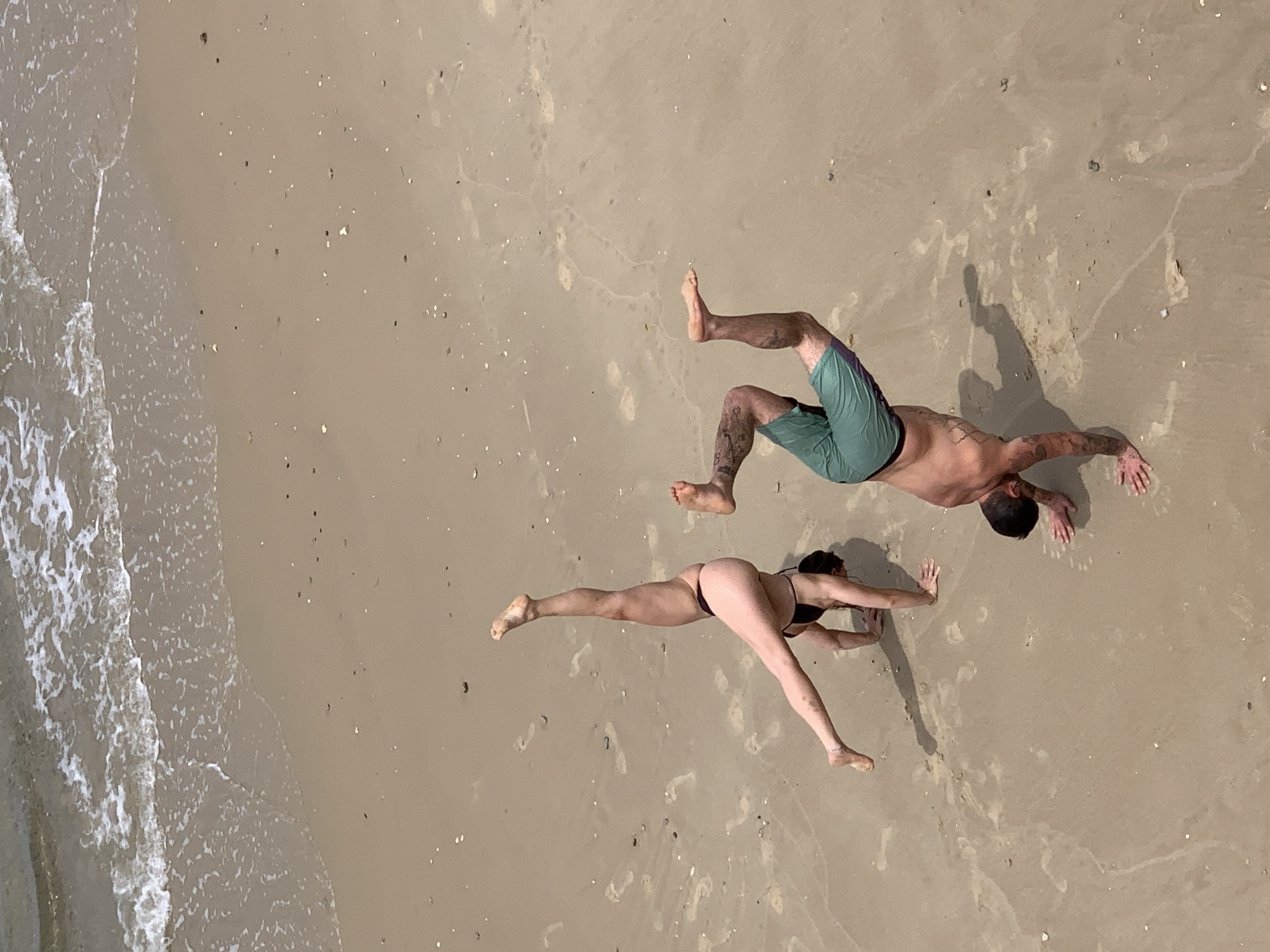 Two individuals are positioned on a sandy beach adjacent to an ocean coastline. One person wears green shorts, while the other is in a dark-colored bikini. Their bodies are in positions suggesting either movement or dance on the sand. The setting appears to be outdoors. It is not possible to confirm the location in the city of Tel-Aviv, Israel, based on the image.