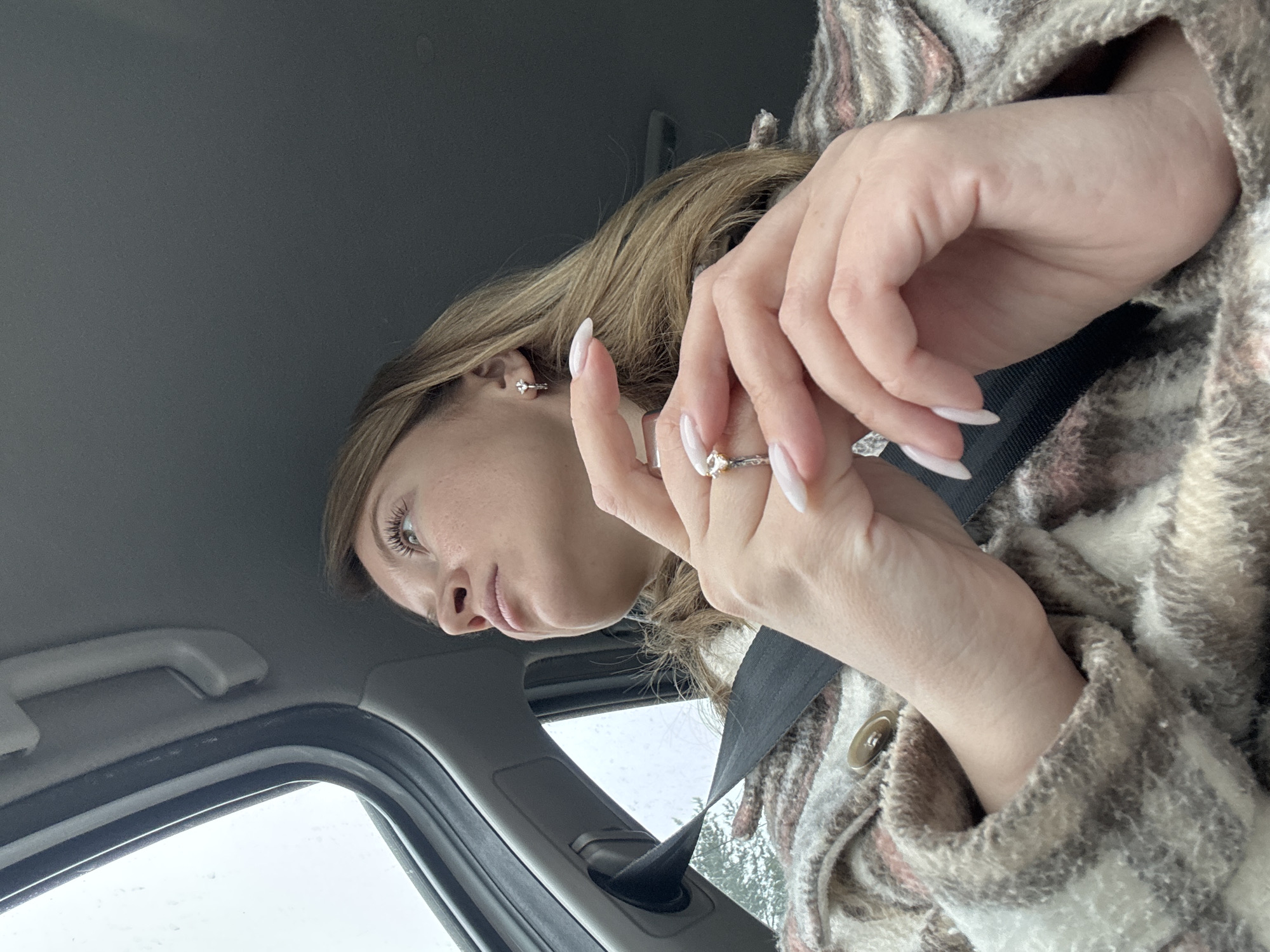 A woman is sitting in a car, wearing a seatbelt and a brown and white patterned jacket. She has light brown hair, earrings, and long white fingernails. She is wearing a ring on her finger and is holding her hands up. The weather appears to be overcast as seen through the car window.
It is not possible to determine the location as Votkinsk, Russia from the given image.