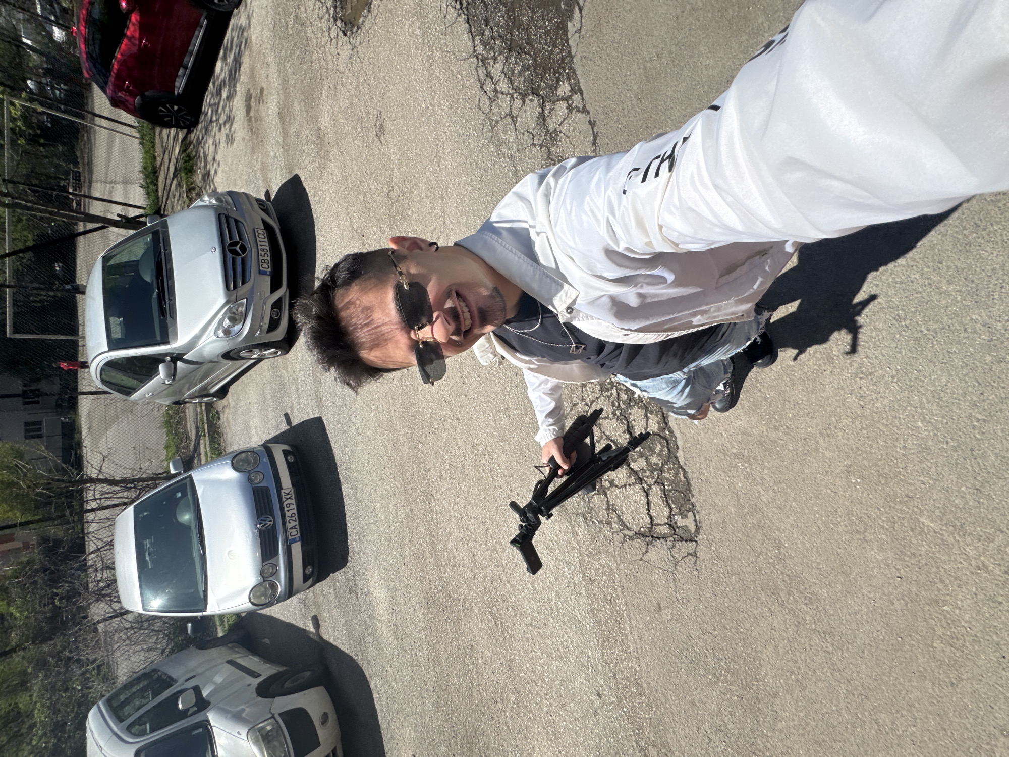 A man stands on a cracked asphalt surface, holding what appears to be a black assault rifle. He is wearing sunglasses, a white jacket, a dark t-shirt, and blue jeans. Three cars are visible in the background: a red hatchback, a silver Mercedes, and a white Volkswagen. The location is likely Sofia, Bulgaria, based on the license plates of the vehicles. A chain-link fence and some sparse vegetation are also present in the background.