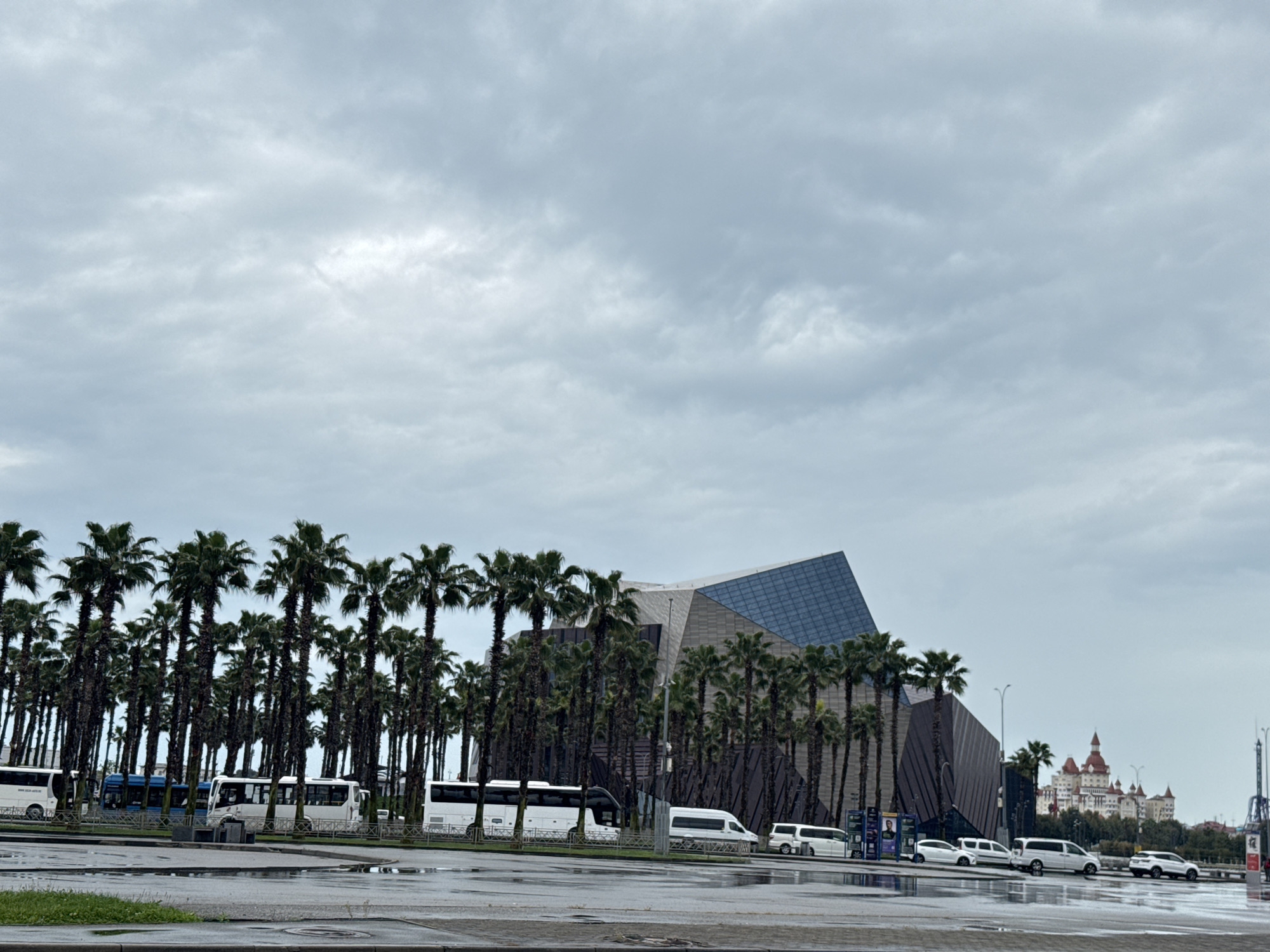 The image depicts a cityscape in Sirius, Russia, under a cloudy sky. In the middle ground, a modern architectural structure with a prominent geometric design featuring glass and dark paneling stands amidst a dense row of palm trees. A line of various white vehicles, including vans and buses, is parked along the road adjacent to the trees. The wet pavement reflects the sky and the surrounding objects. In the background to the right, a fairytale-like castle structure with multiple spires is visible. The overall scene suggests a modern area with a touch of fantasy architecture.
