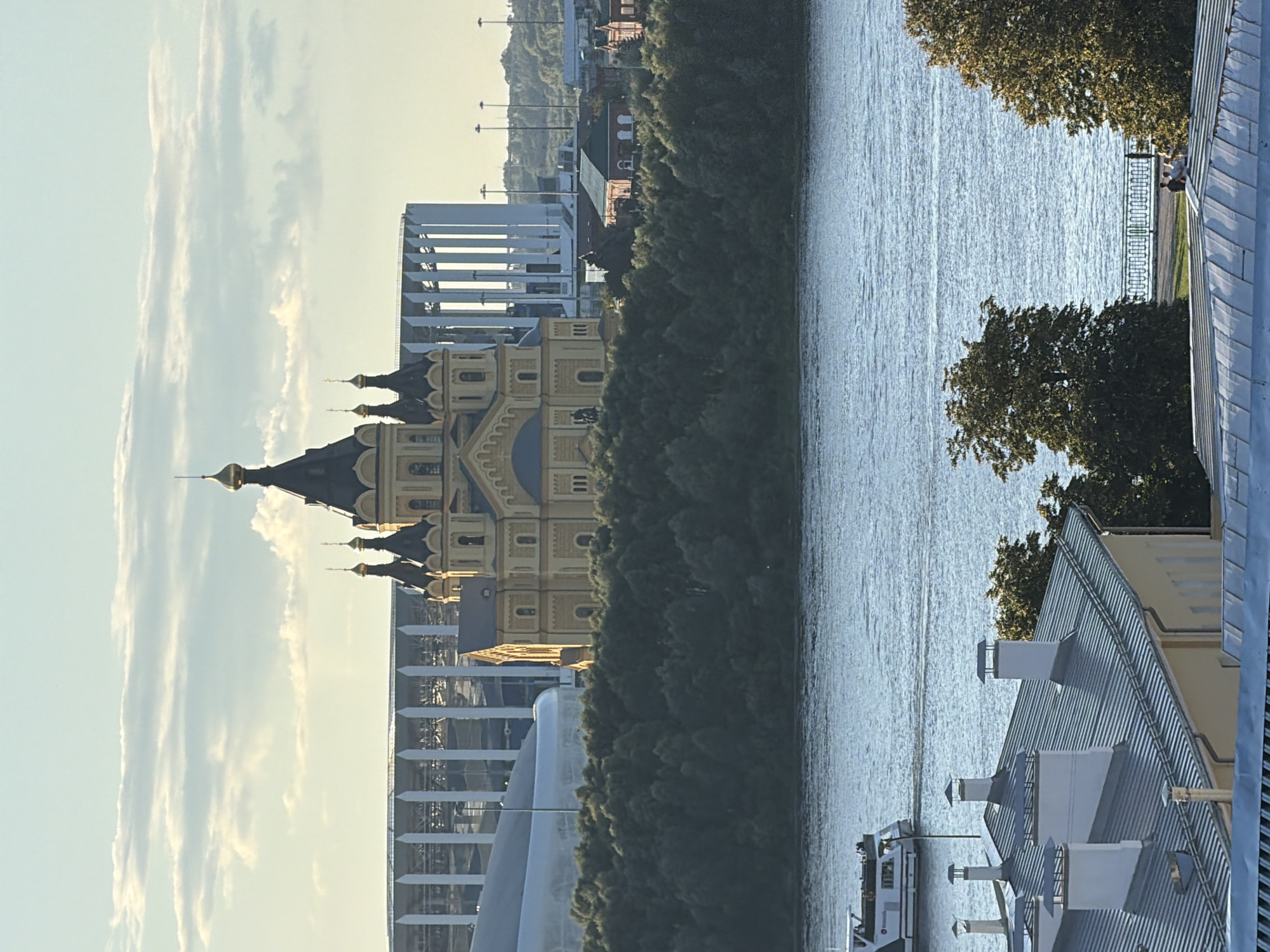 The image shows a view of Nizhny Novgorod, Russia, with a river in the foreground. On the far bank, a yellow orthodox cathedral with multiple domes is visible, situated on a hill covered with trees. Behind the cathedral, a large modern stadium with white pillars and a curved roof is present. In the foreground, several buildings with pitched roofs and chimneys are depicted. A white boat is traveling on the river. The sky is cloudy with patches of blue.