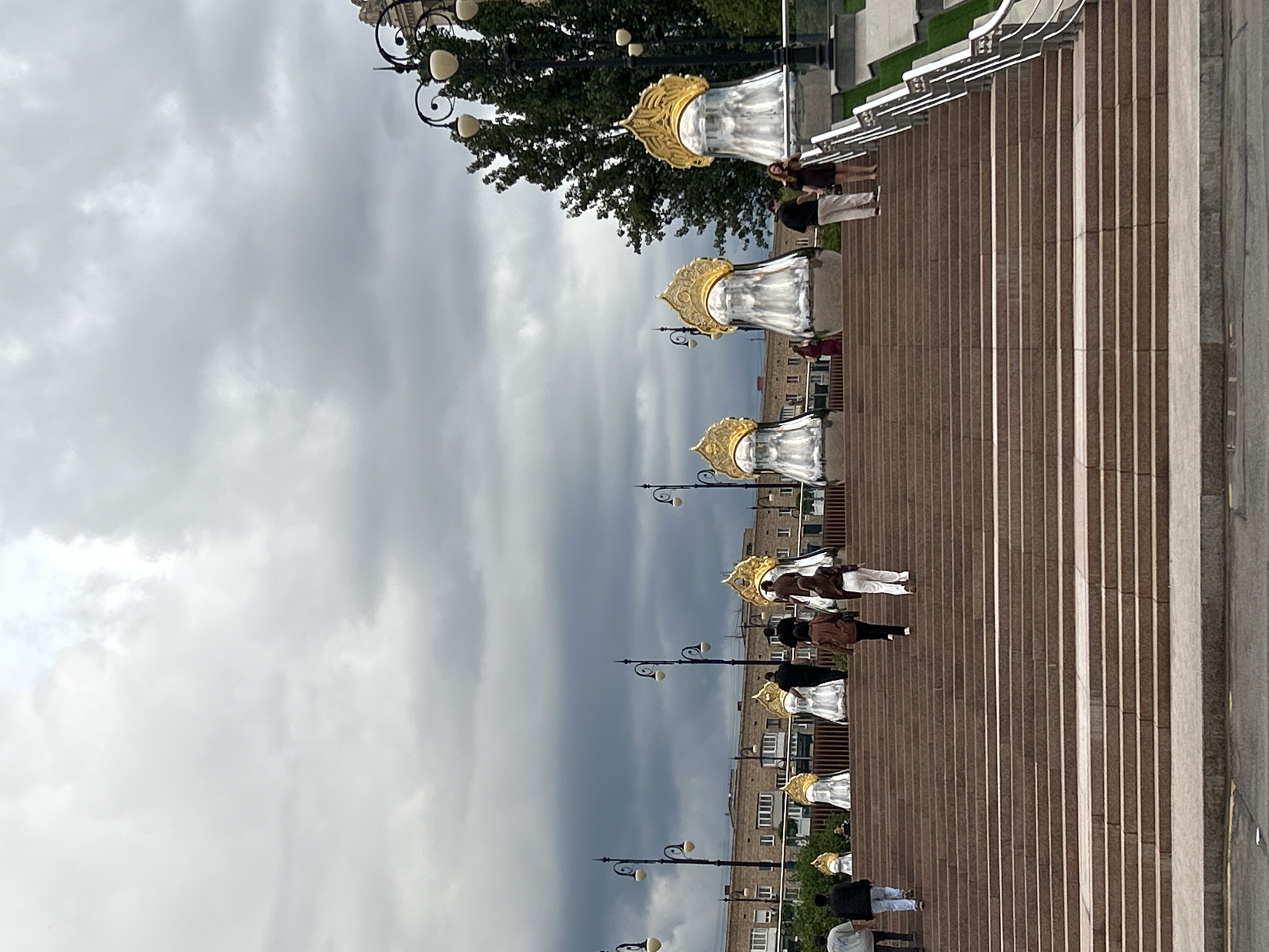 This image captures an outdoor scene on a cloudy day, featuring a wide, grand staircase made of pinkish-brown stone. The staircase is lined with several large, silver sculptures that resemble stylized figures adorned with golden headdresses. Several people are seen ascending the stairs, dressed in casual attire. In the background, there are brick buildings and trees, with lampposts featuring multiple lights visible along the upper portions of the staircase. The overall setting suggests a public urban space, possibly a park or a monument area in Moscow, Russia.