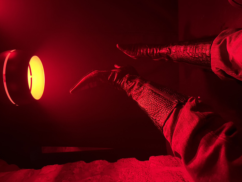 Two legs clad in reptile-patterned boots and denim jeans extend into a darkened, red-lit space. The pointed toe of one boot is close to a cylindrical wall sconce emitting a yellow light. The floor beneath is indistinct but textured, possibly sand or a similar granular surface, also bathed in red. The overall ambiance is mysterious and dramatic due to the stark contrast and limited visibility.