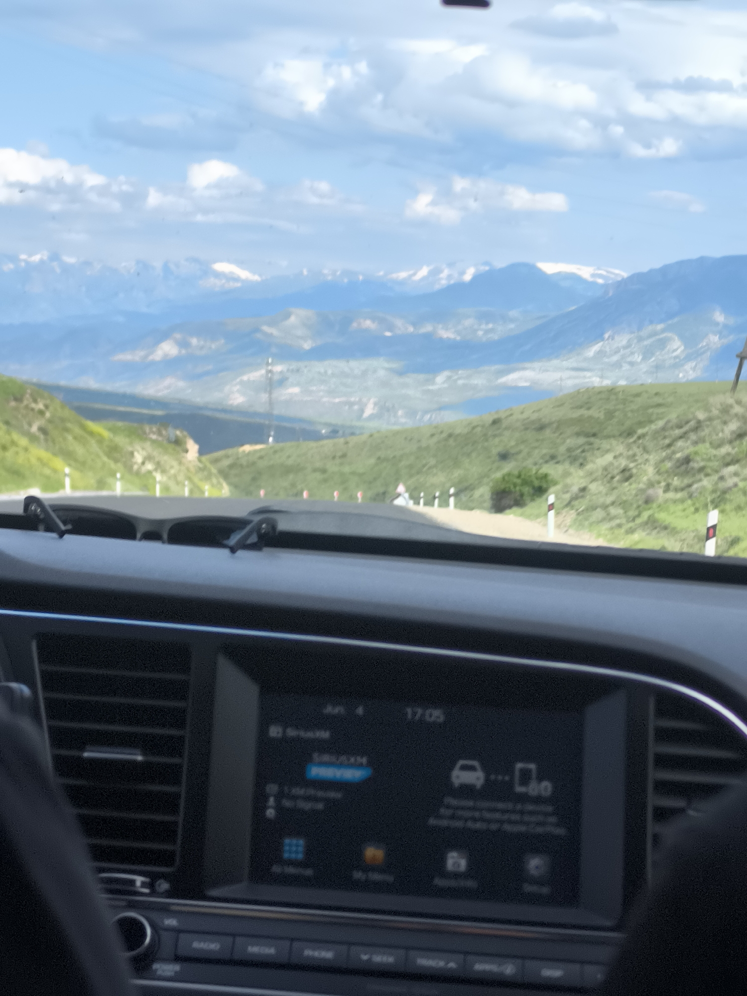 The image presents a view from inside a vehicle, looking forward onto a mountainous landscape in Yelpin, Armenia. The foreground is dominated by the car's dashboard, featuring air conditioning vents on the left and a digital infotainment screen in the center.
On the car's screen, the time "17:05" is clearly displayed. Below this, "SiriusXM PREVIEW" is visible, along with a partial date "Jun 4". Various icons are present, including a car symbol and a phone symbol, below which some smaller, blurry text appears, likely instructions related to connecting a device. Physical buttons below the screen are labeled "RADIO", "MEDIA", "PHONE", "SEEK", "TRACK", "APPS", and "SETUP". Parts of a person's hands are visible at the bottom left and right edges, suggesting occupants are present.
The external scene depicts a road winding through green, rolling hills. White and red/black striped road markers line the edge of the asphalt. In the middle distance, the landscape transitions to larger mountains, covered in green vegetation. A faint structure, possibly a utility pole, can be seen in the valley. The background features a vast expanse of snow-capped mountains under a bright blue sky with scattered white clouds. The presence of snow on distant peaks, combined with the green foreground, suggests a spring or early summer season. The fair weather and clear visibility highlight the extensive mountainous terrain.