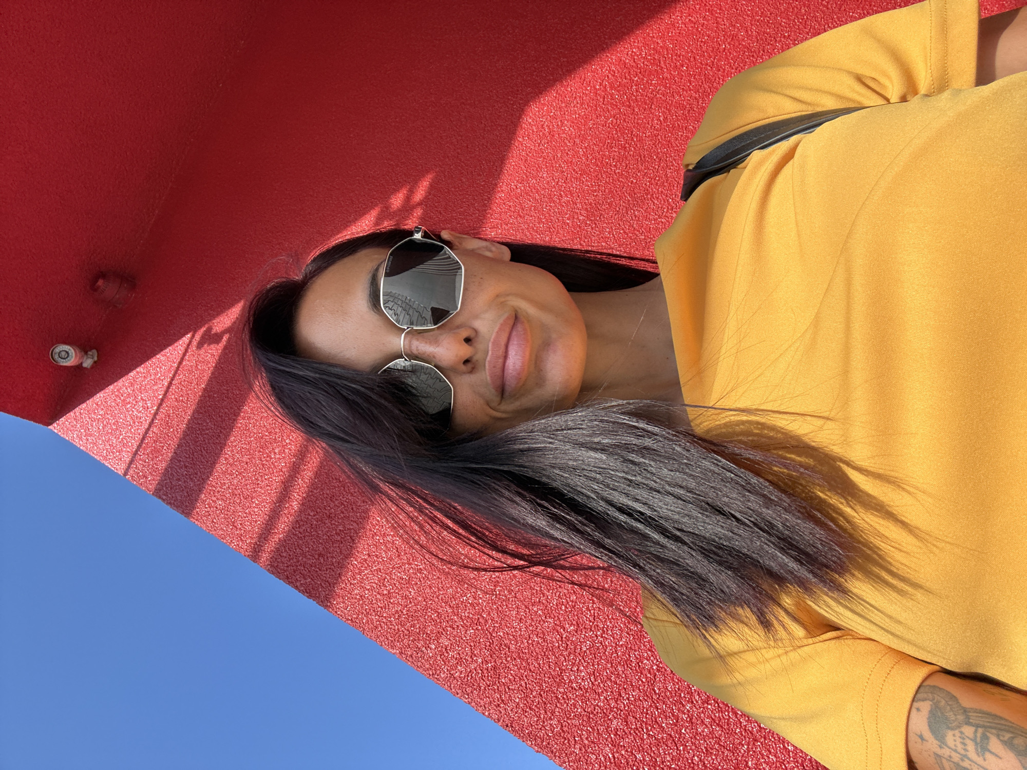 A low-angle, medium shot captures a woman with long, dark hair and sunglasses against a bright red wall and a clear blue sky. She is wearing a bright yellow t-shirt. A tattoo is visible on her left arm. The red wall features shadows and what appear to be two security cameras. The woman is looking slightly off-camera and smiling. The image does not contain enough information to determine the location within the city of Pleven, Bulgaria.