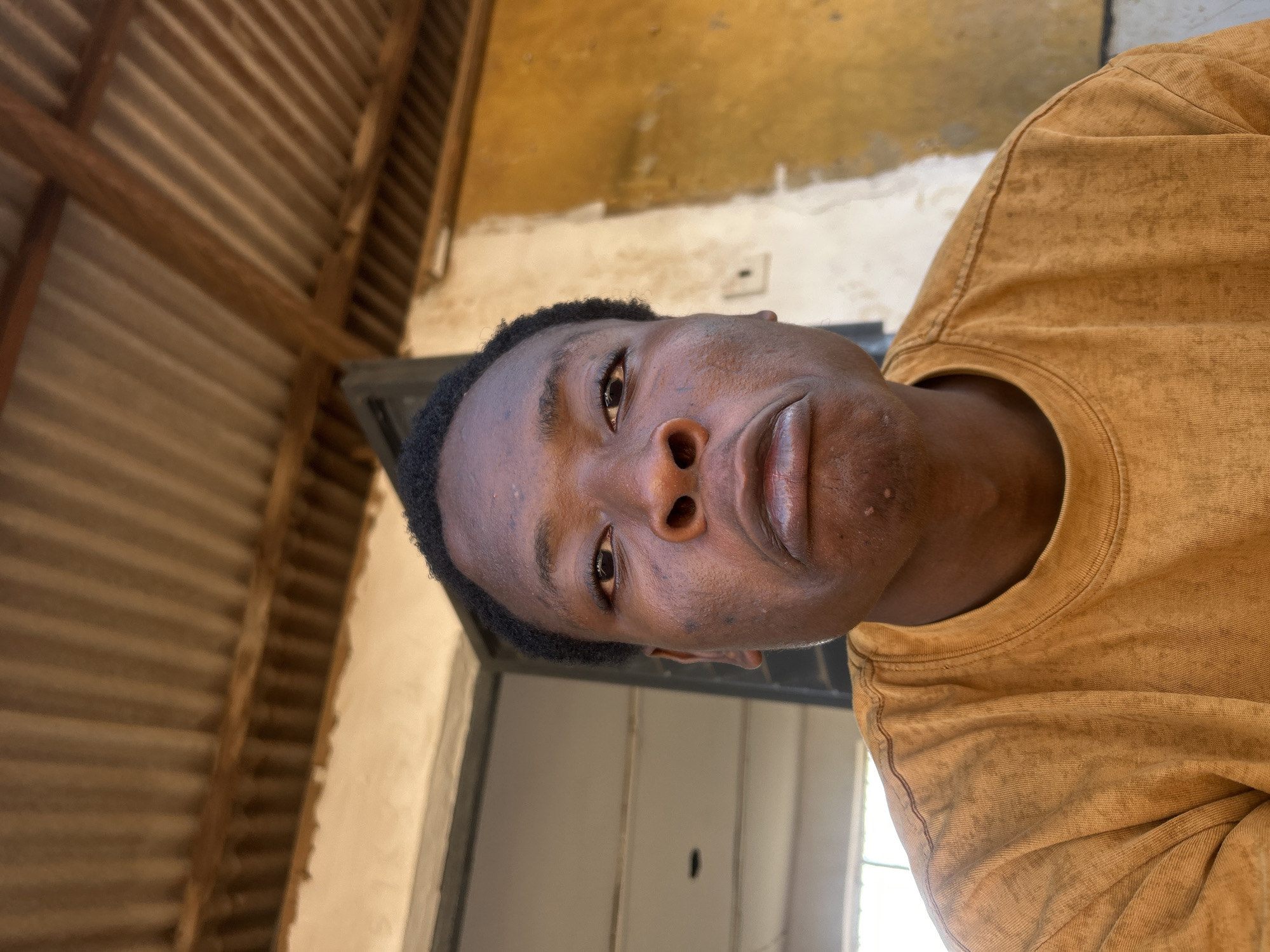 A medium close-up portrait shows a young man with dark skin and short, dark hair looking directly at the camera. His head is tilted significantly to the right, suggesting the photo was taken at an angle or the subject is leaning. He wears a mustard yellow or faded brownish short-sleeved t-shirt with a subtly textured or washed appearance. His expression is neutral to slightly serious, with his lips gently pursed.
The background is simple and appears to be an indoor or covered outdoor space. On the left side, a slatted wooden structure, likely a ceiling or roof support, is visible. Above and to the right of the subject, a wall with uneven, yellowish-ochre paint or plaster suggests a weathered or unfinished surface. Further down on the right, behind the subject's right shoulder, there's a lighter wall section and what might be an open doorway or window. The scene is brightly lit, indicating it is daytime. There is no visible text in the image.