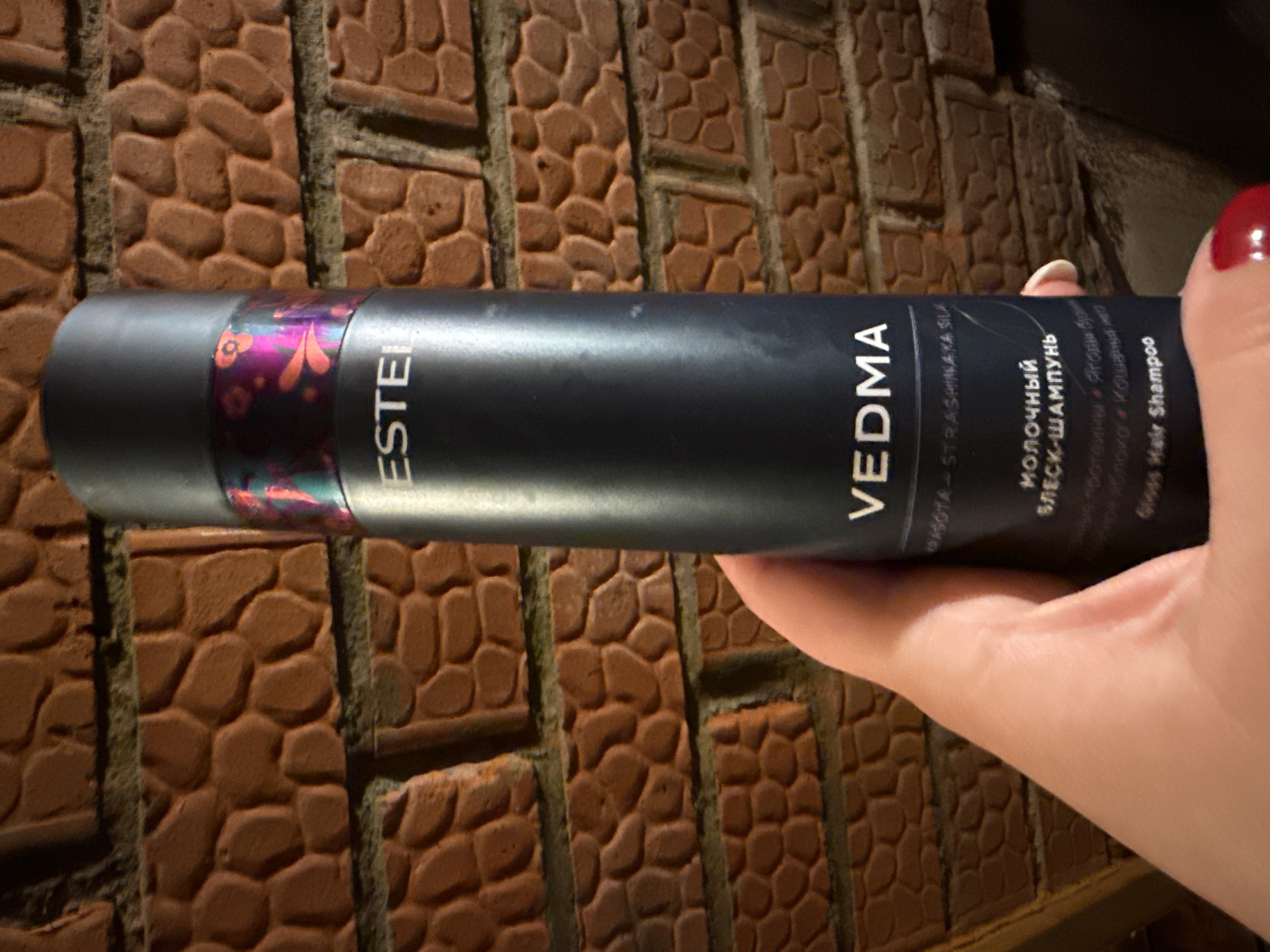 A hand holds a gray cylinder of ESTEL VEDMA gloss hair shampoo in front of a textured brown brick wall. The shampoo bottle's label is partially visible and includes text in Cyrillic and English. The bottle features a colorful decorative band near one end. A part of the hand and a thumbnail with red nail polish are visible. The location cannot be determined from the given information, nor is any city mentioned.