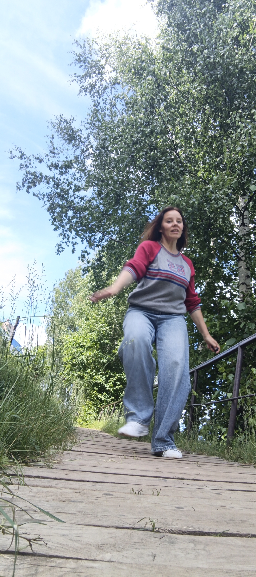 A young woman with brown hair is captured mid-jump on a wooden walkway in Chaikovsky, Russia. She is wearing a gray long-sleeved shirt with red sleeves and blue and red stripes across the chest, and loose-fitting blue jeans. Her white sneakers are visible as she is in motion. The walkway is made of weathered wooden planks and is surrounded by lush green grass and trees. The sky above is a bright blue with some white clouds. A metal railing is visible to the right of the walkway, partially obscured by foliage. The scene appears to be an outdoor recreational area.