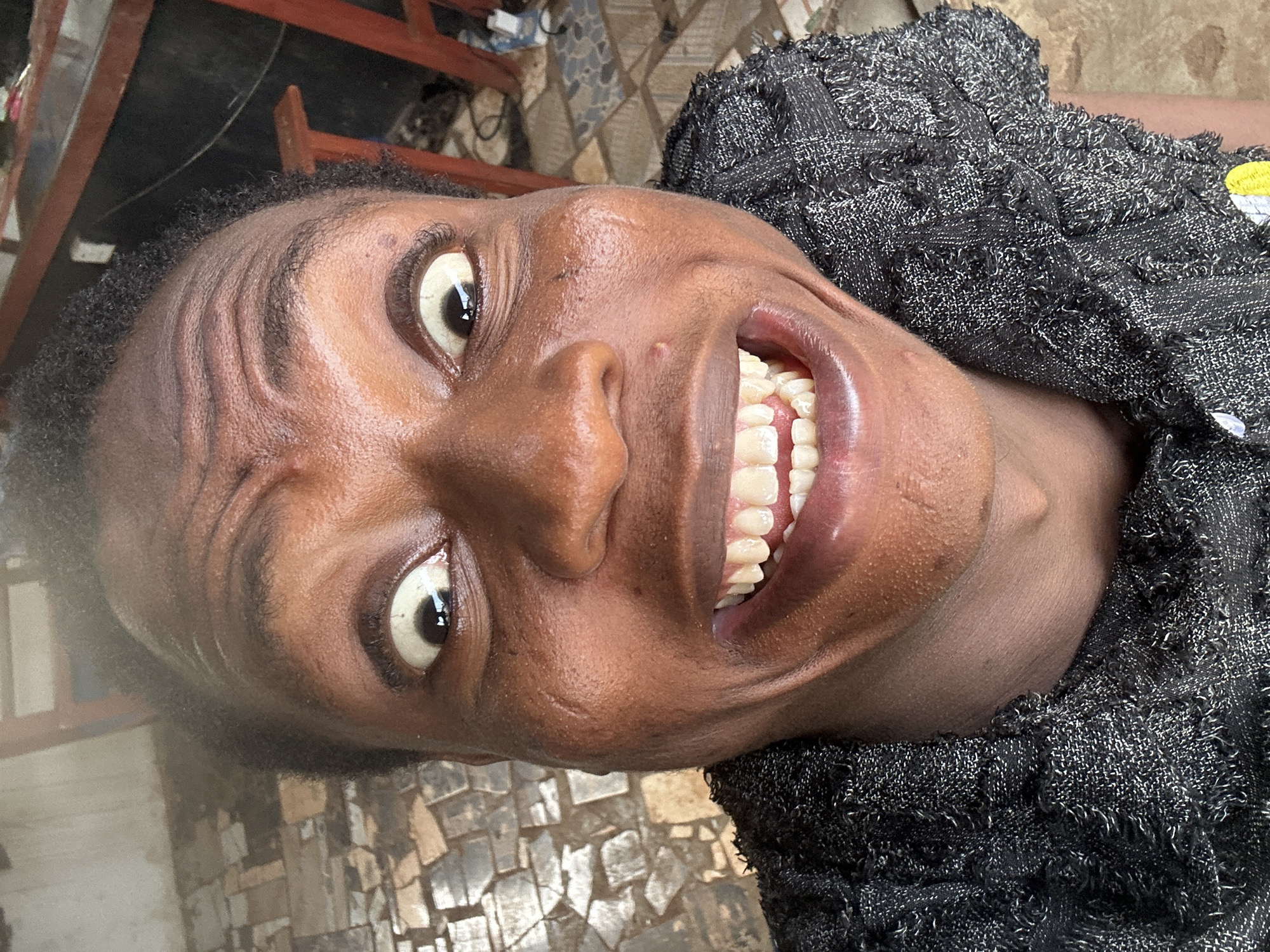 A close-up, angled shot of a dark-skinned person, presumably female, with a wide smile revealing straight, white teeth. The individual is wearing a textured black garment. The background is indistinct but shows a tiled or stone-patterned surface. The location is noted as Unknown, Unknown.