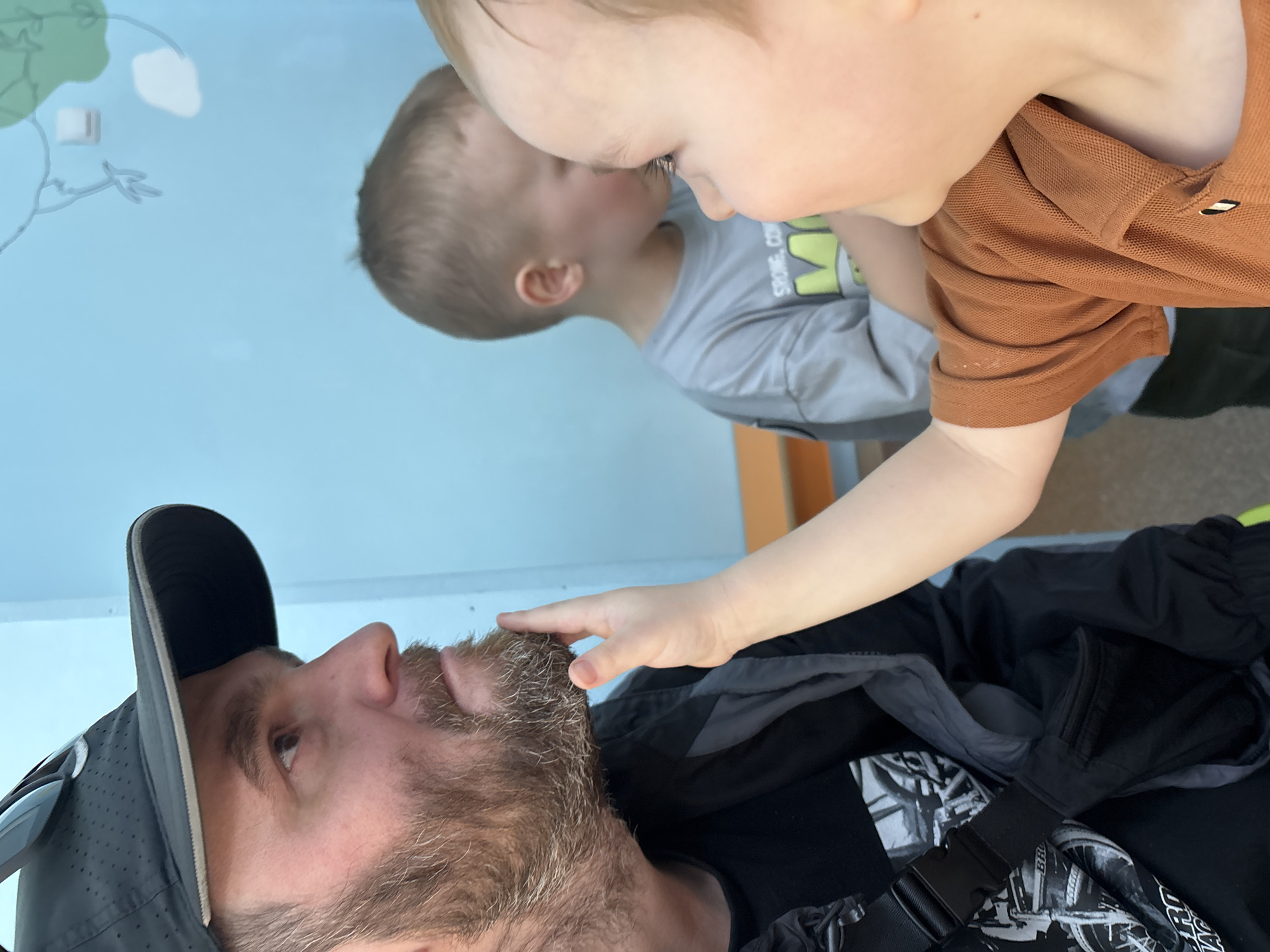 The image shows a man wearing a black baseball cap and a black jacket, with two young children. One child is wearing a brown polo shirt and is touching the man's beard. Another child is wearing a grey t-shirt and has his face very close to the child in the brown shirt. The background is a light blue wall with a drawing of a tree. The location is most likely indoors, possibly at a children's museum, but the setting is unclear. There is no information to indicate this photo was taken in the city of Chaikovsky, Russia.