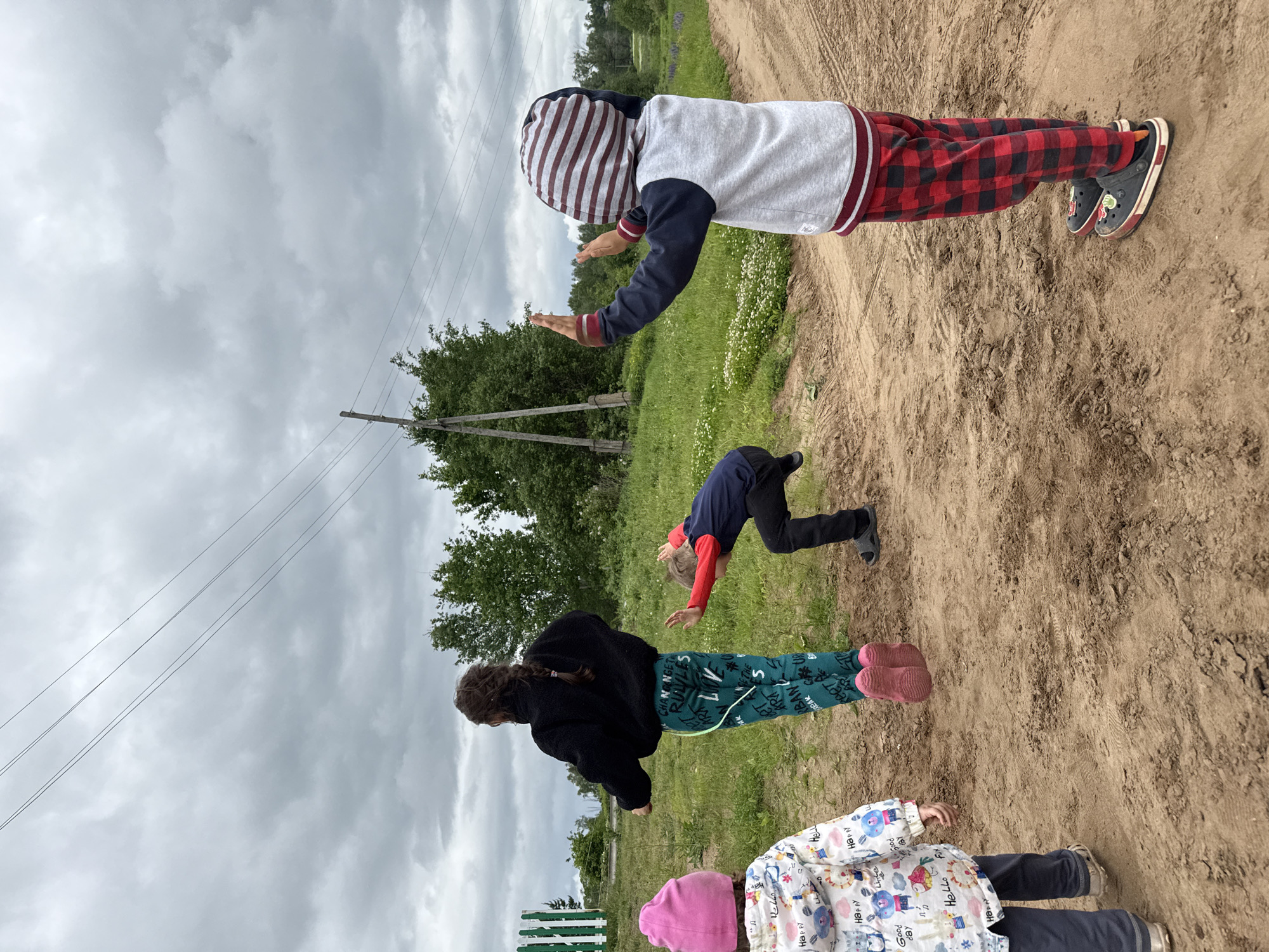 The image displays four children on a dirt path in a rural area of Уральское, Russia, under an overcast sky. From left to right: a small child is visible from the rear, wearing a pink knit hat, a white patterned jacket with various English text including "Happy Good day" and "Hello," dark pants, and gold-colored shoes. Next, an older child, presumed female, is seen from behind with dark hair in a braid, wearing a black fleece jacket and teal pants with white lettering. This child appears to be in motion, possibly mid-jump or landing, wearing pink footwear. To the right, a boy is positioned low to the ground, leaning forward with arms outstretched, wearing a dark blue and red long-sleeve shirt, black pants, and dark sandals. The fourth child, an older boy, stands further right, viewed from the rear, wearing a grey and navy hooded jacket with a red and white striped hood, red and black plaid pants, and dark Crocs adorned with red cherry-like designs. He has both hands raised with palms forward, performing a gesture. The dirt path shows visible tire tracks and footprints. Green grass and small white wildflowers flank the path, with patches of purple flowers further in the background on the right. A wooden utility pole with multiple power lines is prominent in the mid-ground on the right, extending across the upper portion of the frame. A green and white striped wooden fence is visible on the far left. Dense foliage and trees line the horizon under the uniformly grey, cloudy sky.