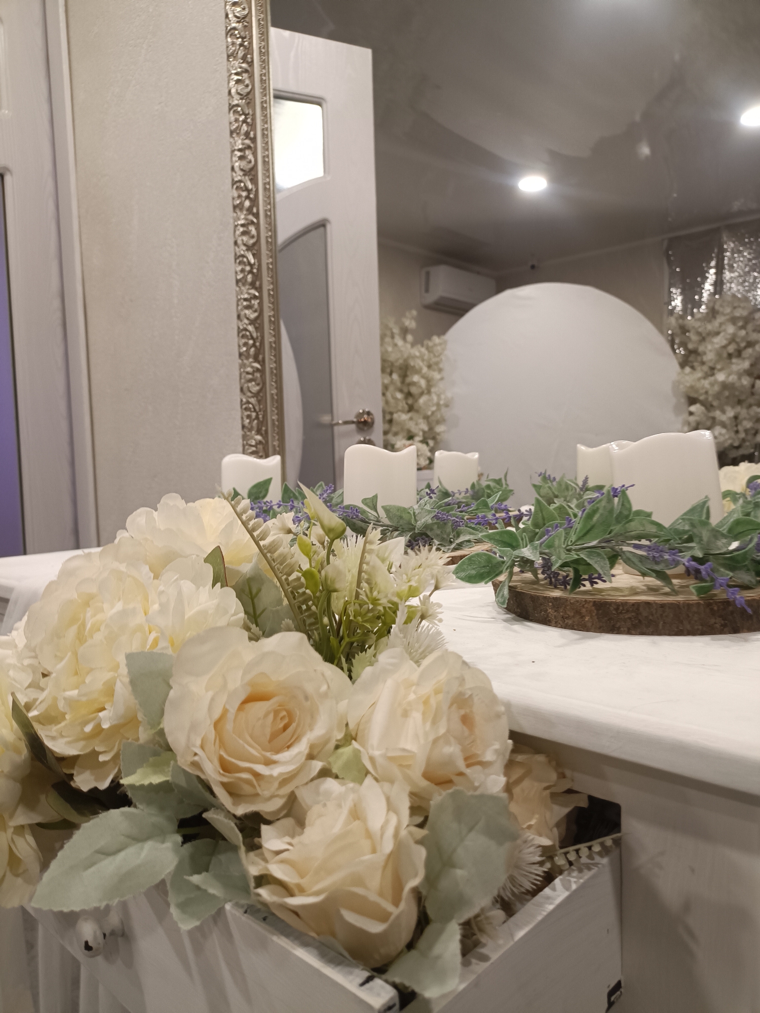 The image captures an elegantly decorated indoor setting, likely an event space. In the foreground, a lavish floral arrangement consisting of numerous light cream or off-white artificial peonies and roses, complemented by soft green foliage, is displayed in a white wooden container, possibly a drawer integrated into a table.
Behind this, in the midground, a centerpiece is visible on a white surface, which appears to be a table or counter. This centerpiece features several white pillar candles, some with gently wavy tops, arranged on round slices of natural wood. These candles are surrounded by artificial lavender sprigs with purple accents and lush green leaves.
The background reveals more of the sophisticated environment. To the left, a white door with an ornate, silver-colored decorative trim on its frame is prominent. Further back and to the right, a large, smooth white spherical or dome-like structure stands, surrounded by delicate white artificial trees resembling cherry blossoms. The entire background area, including the dome, trees, and the ceiling with recessed lighting and an air conditioning unit, is reflected in a large, shiny surface or mirror on the right side of the image, creating an illusion of expanded space. A subtle hint of a silver sequined curtain is also visible in the reflected background.
The overall decor, with its light color palette, floral arrangements, and candles, suggests a celebratory event such as a wedding reception, a party, or a decorative display for a special occasion. The setting appears to be indoors with artificial lighting, and there is no visible text in the image.