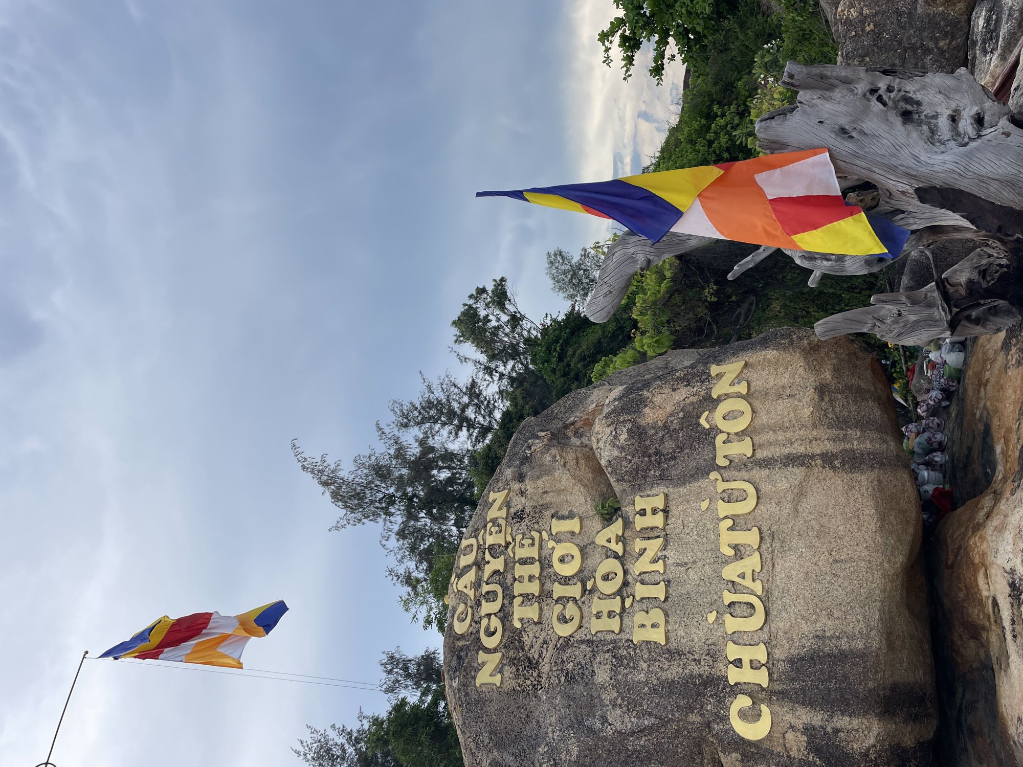 The image captures a large, weathered rock face adorned with large, golden, raised lettering. The inscription reads "CẦU NGUYỆN THẾ GIỚI HÒA BÌNH CHÙA TỪ TÔN" which translates to "Pray for World Peace Tu Ton Pagoda". Two Buddhist flags are visible. One is a traditional Buddhist flag with six vertical stripes of blue, yellow, red, white, orange, and blue, flying from a pole extending from a structure to the left. The other is a more contemporary Buddhist flag, also composed of multiple colored panels, affixed to a weathered, grey, driftwood-like structure in the foreground. The background features lush green trees and a cloudy sky, suggesting an overcast day. The location is identified as the Tu Ton Pagoda in Nha Trang, Vietnam, known for its stunning coastal scenery and religious significance, perched on a rocky outcrop overlooking the sea. The area around the base of the rock appears to be a small shrine, with various offerings visible, including white and colored bags, and small figurines. The overall impression is one of tranquility and spiritual devotion.