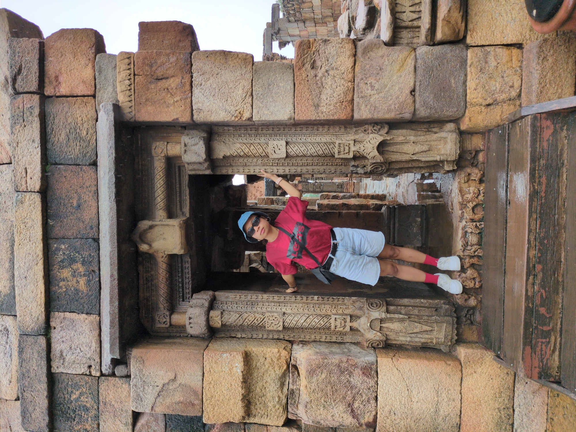 A young woman wearing a red t-shirt, denim shorts, a blue bucket hat, and white sneakers stands in a stone archway at Qutub Minar in Delhi, India. She is leaning against a carved stone pillar and looking towards the camera. The archway is made of rough-hewn stone blocks, and the ground in front of it is wet, suggesting recent rain. The background shows more stone structures and ruins.