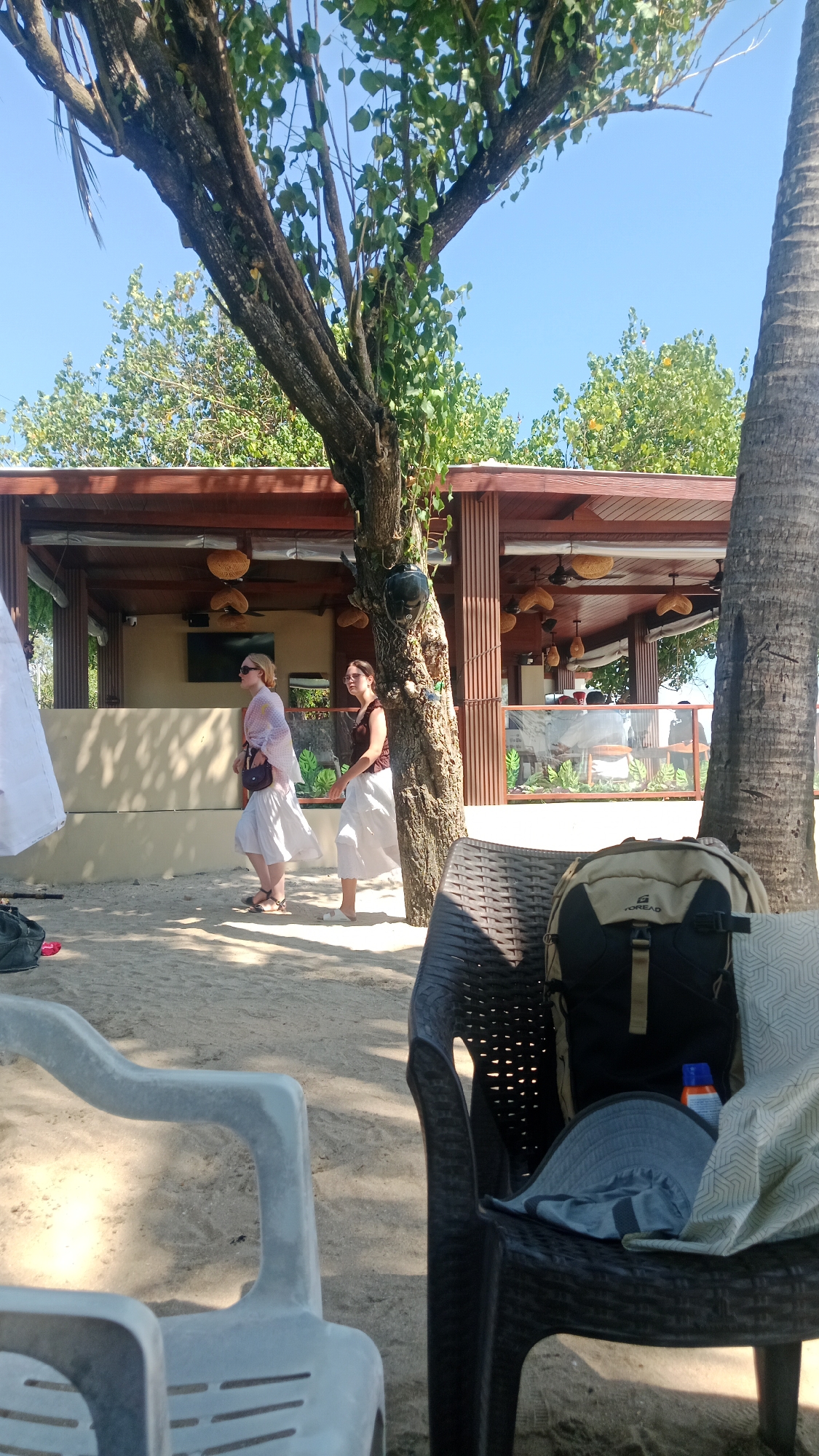 Two women are walking on a sandy beach in Kuta, Indonesia, near an open-air restaurant. The woman on the left is wearing a light pink translucent shawl over a white skirt and sandals, with a dark purse slung over her shoulder. The woman on the right is wearing a brown top and a white skirt, also in sandals. They are walking away from the viewer and towards the restaurant, which has a thatched roof and wooden pillars. The restaurant appears to be situated behind a low wall and has glass partitions. In the foreground, two chairs are visible: a light gray plastic chair and a black rattan chair. A backpack and a hat are placed on the black chair. Trees, including a palm tree on the right, are present in the scene, casting shadows on the sand. The sky is clear and blue. The overall scene suggests a relaxed atmosphere on a sunny day at a beachside establishment.