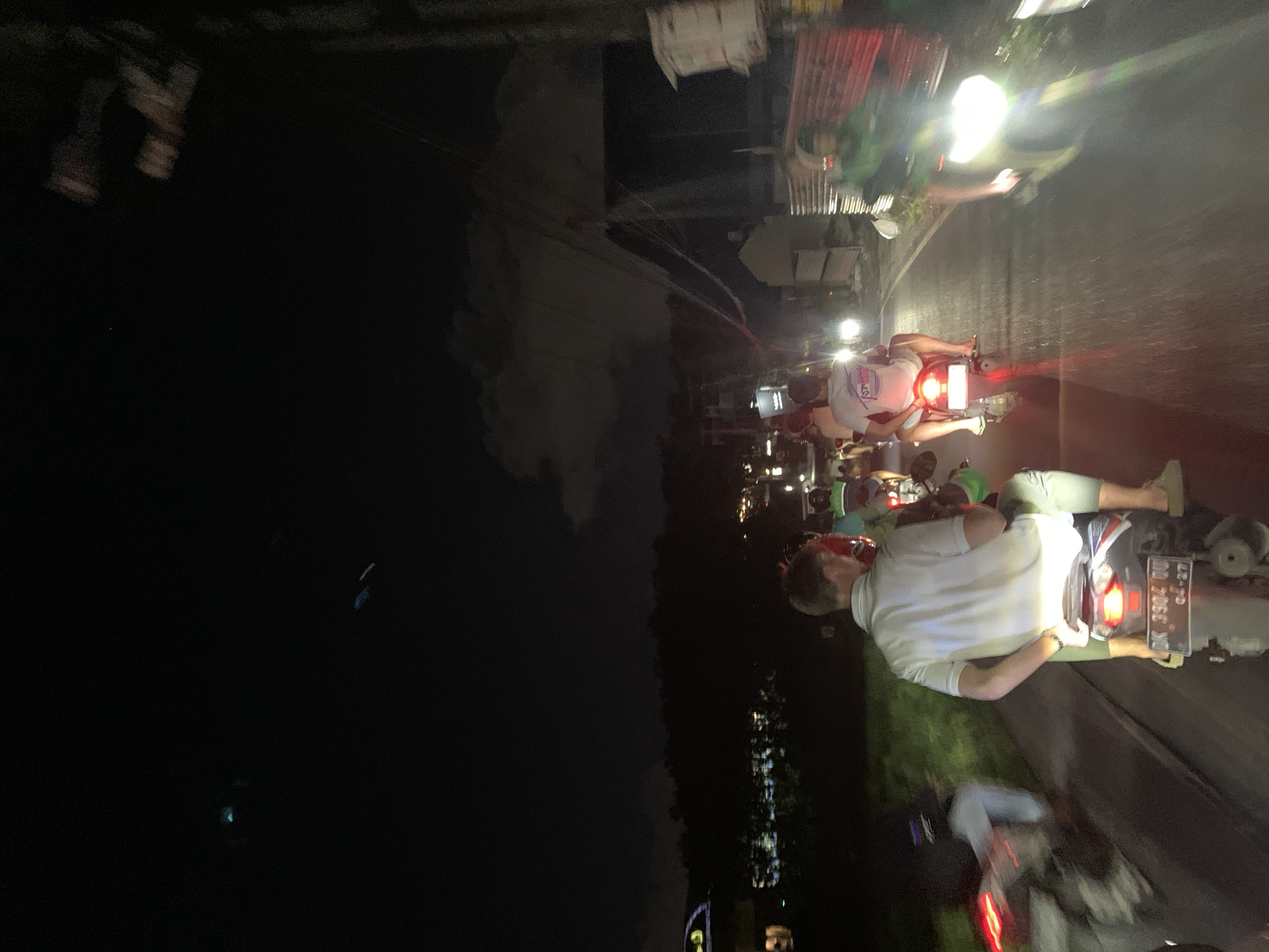 A nighttime photograph shows a group of people riding motorcycles on a road in North Kuta, Indonesia. The motorcycles have their headlights and tail lights on. The riders are wearing casual clothing, and some are wearing helmets. The surrounding environment is dark, with buildings and trees visible in the background.