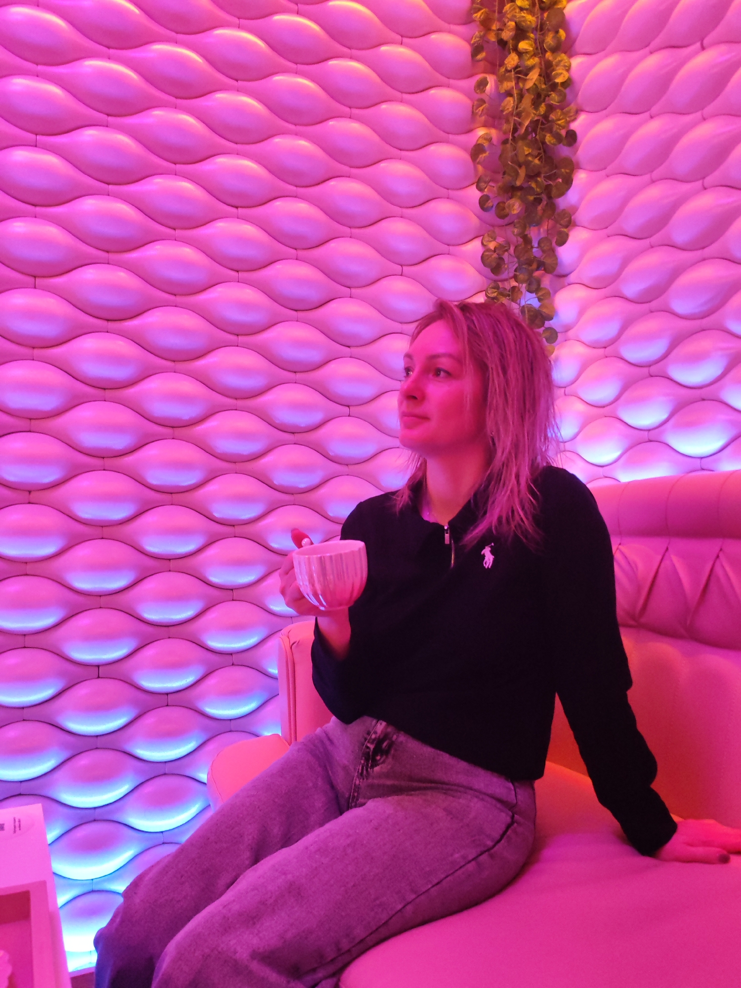 A woman is seated on a pink cushioned bench in a room with unique wall decorations, holding a small cup. The room is illuminated with a pink and blue light. The walls have a textured, wavy pattern, and a decorative vine hangs from the top corner of the wall. The woman is wearing a black long-sleeved polo shirt and light-colored denim pants. The scene is captured in what appears to be an indoor setting with the light suggesting a particular atmosphere or mood, possibly in a cafe or lounge with the wall pattern reflecting a particular style of interior design. There is a small table on the left with a white base. This image was taken in Izhevsk, Russia.