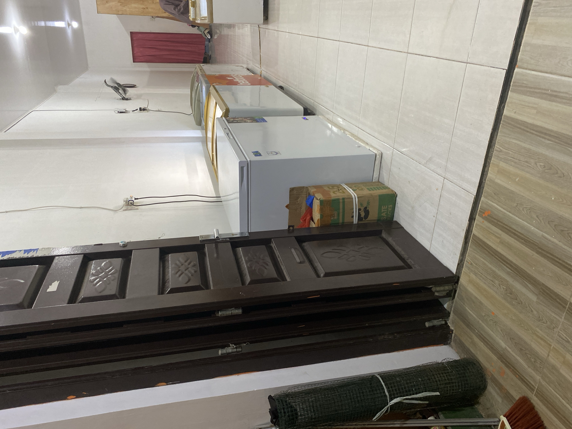 The image depicts an indoor scene, likely a commercial establishment, with elements suggesting it is located in Payatieng, Indonesia. In the foreground, to the left, is a brown wooden door propped open, revealing a glimpse of its interior paneling with decorative carvings. A roll of dark green netting, bound with white ties, stands upright next to the door. A broom with an orange brush lies on the wooden floor in the lower left corner.
The main area of the scene is dominated by several chest freezers lined up against a white wall. The freezers are varied in color, with one being predominantly white, and others featuring yellow and orange accents. Boxes are stacked near the front of the white freezer. Electrical cords are visible plugged into a wall outlet. A black wall-mounted fan is positioned higher on the white wall. Further down the wall, a red curtain hangs, obscuring a doorway or opening. In the background, on the right, a person is partially visible, seated. The floor throughout the space is tiled, with a transition to wooden flooring where the door is situated. The lighting suggests an interior space with overhead fixtures.