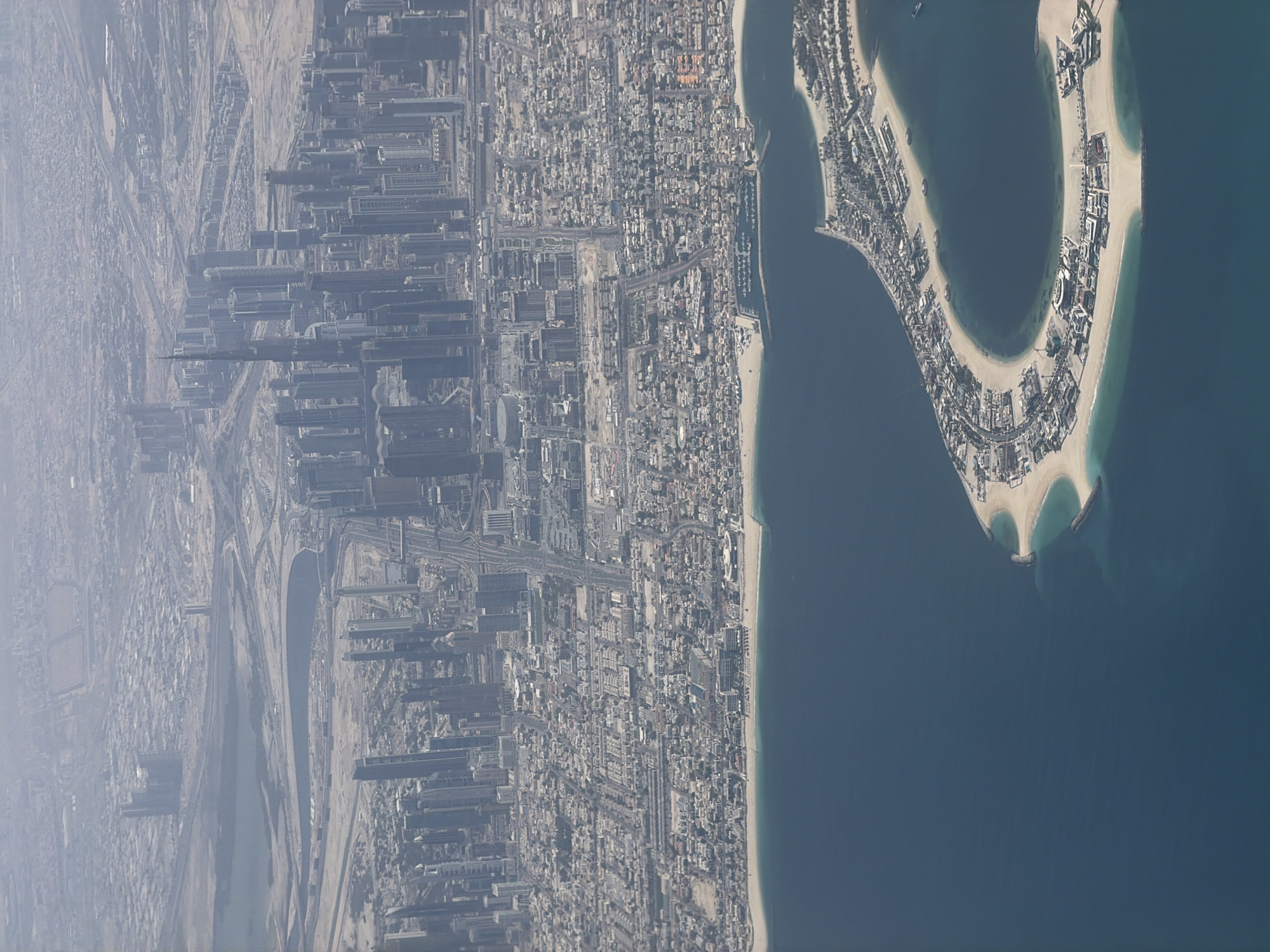 An aerial, slightly rotated image presents a vast urban landscape adjacent to a large body of water. The scene is dominated by a dense cityscape with numerous buildings, including a prominent cluster of tall skyscrapers and an extensive network of residential and commercial structures. Roads and infrastructure crisscross the urban sprawl, which gradually transitions into more undeveloped, sandy, or arid terrain further inland to the left.
The right side of the image features a wide expanse of deep blue water, bordering a light sandy coastline. A striking, crescent-shaped artificial island or peninsula is visible in the water, densely developed with buildings along its curved segments and featuring light sandy beaches. Smaller landforms or breakwaters are also present around the island.
The image appears to be taken during the daytime, under bright but hazy atmospheric conditions, which slightly reduces clarity and distance visibility. No specific human activity or people are discernible due to the high aerial perspective. There is no visible text. The distinctive artificial crescent island and the modern high-rise skyline serve as strong visual cues, characteristic of coastal developments found in places like Dubai.