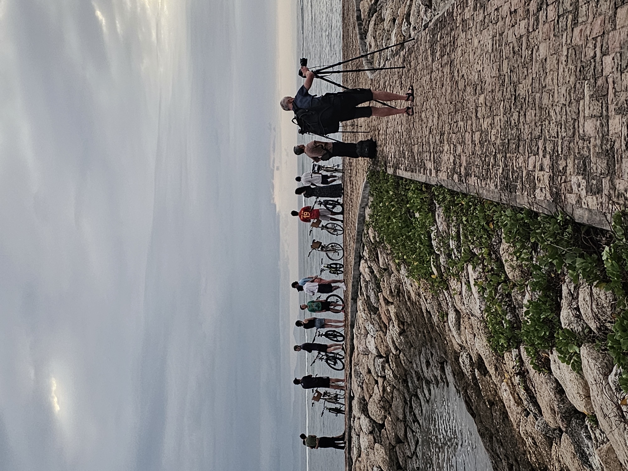 A man takes photos with a tripod, as a line of people with bicycles watch the ocean. The location is Denpasar, Indonesia, on a path lined with paving stones and a stone wall covered in green vegetation. The ocean is visible in the background, along with a cloudy sky.
