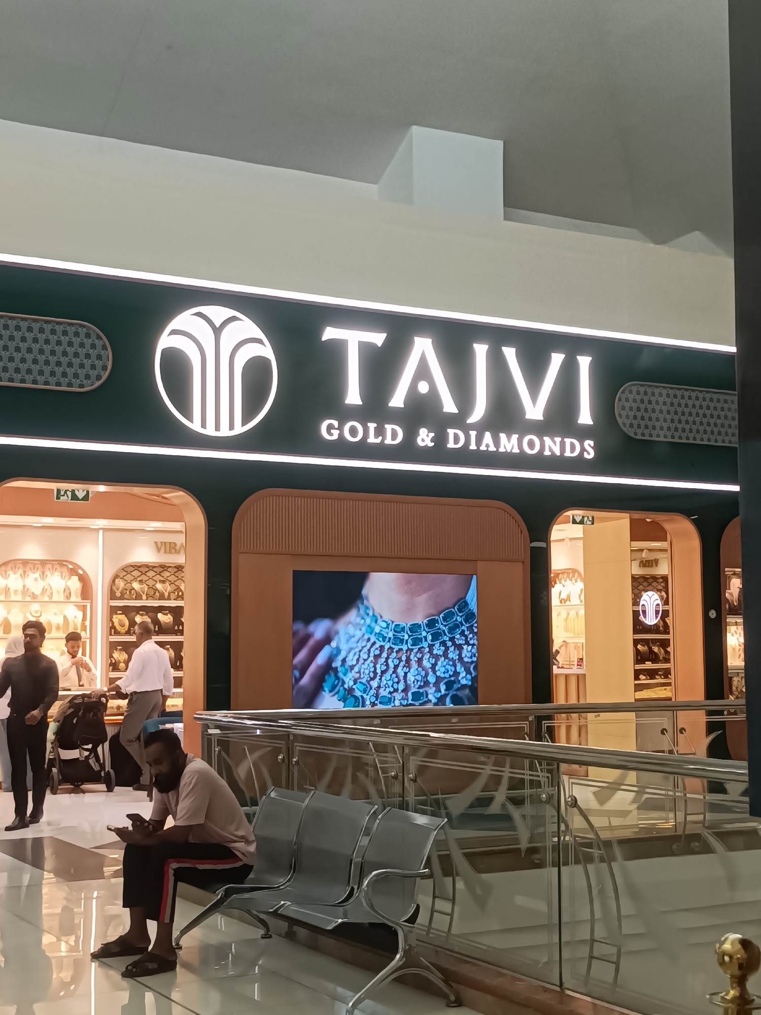 The image shows the storefront of "TAJVI GOLD & DIAMONDS" in Dubai, United Arab Emirates. The store features a large illuminated sign with the brand name and logo. Inside the store, jewelry is displayed on shelves. Several people are visible in and around the store. In the foreground, a man is sitting on a bench, looking at his phone. Another man is walking past the bench. Further back, two men are standing inside the store, and another man is pushing a stroller. The environment appears to be an indoor mall or shopping center, with polished floors and reflective surfaces.